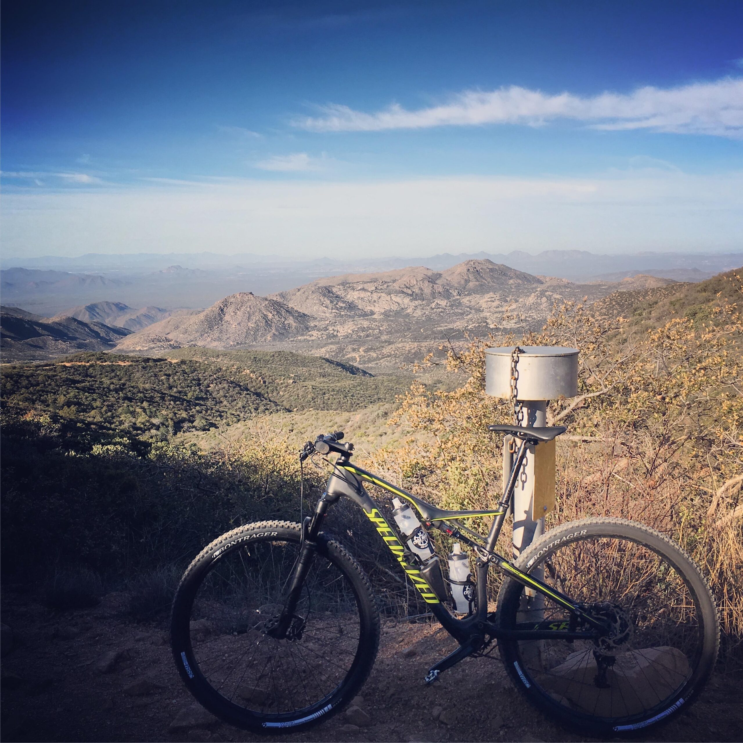 Specialized Epic Expert Carbon World Cup: A mountain bike positioned on a rocky trail overlooking a vast mountainous landscape under a clear blue sky. In the background, rolling hills and distant peaks are visible, while a metal post with a chain stands nearby.