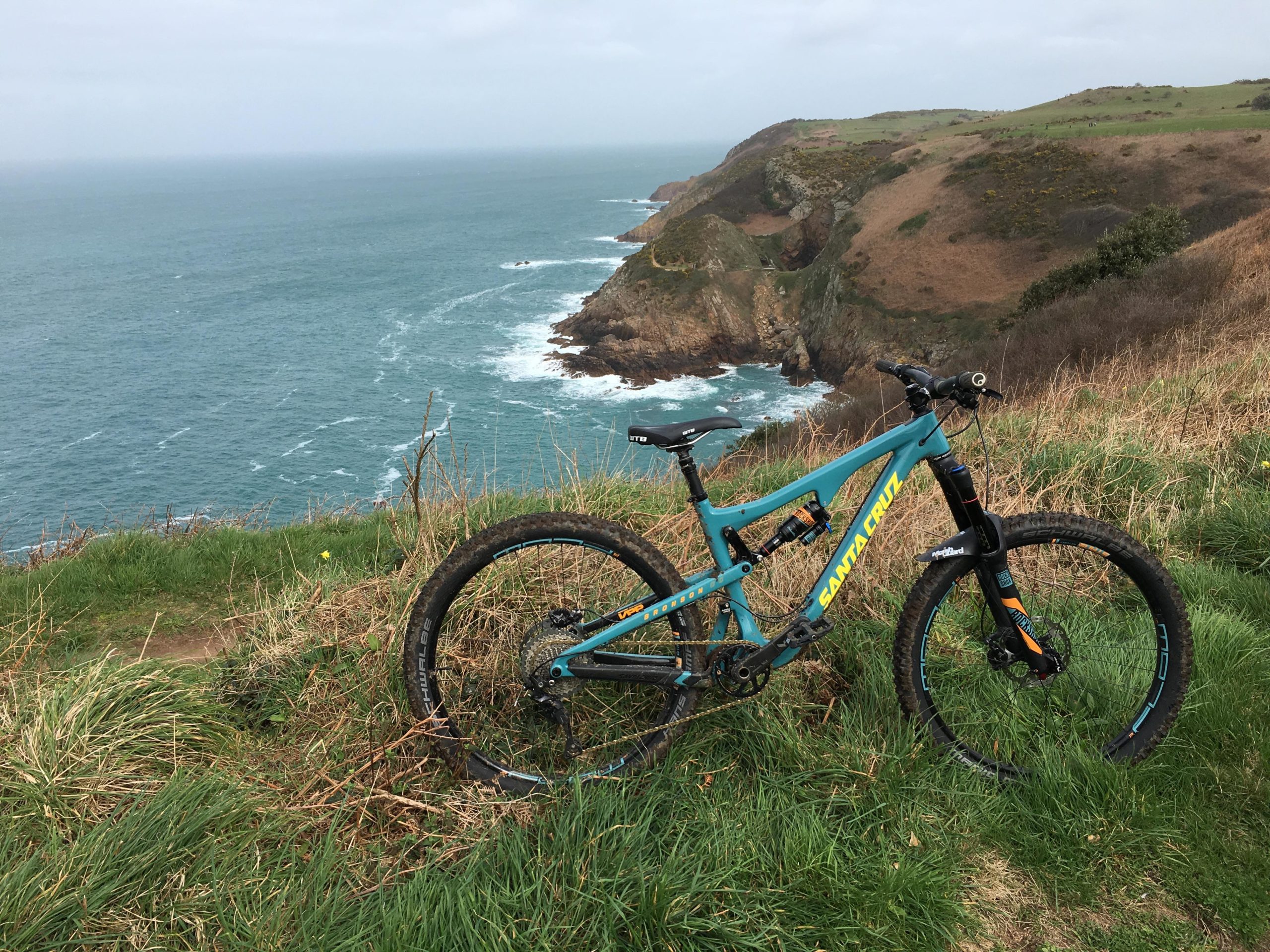 A mountain bike parked on grassy terrain overlooking a rocky coastline and the ocean, with waves crashing against the shore under a cloudy sky. Devils Hole and BACK mountain bike trail.
