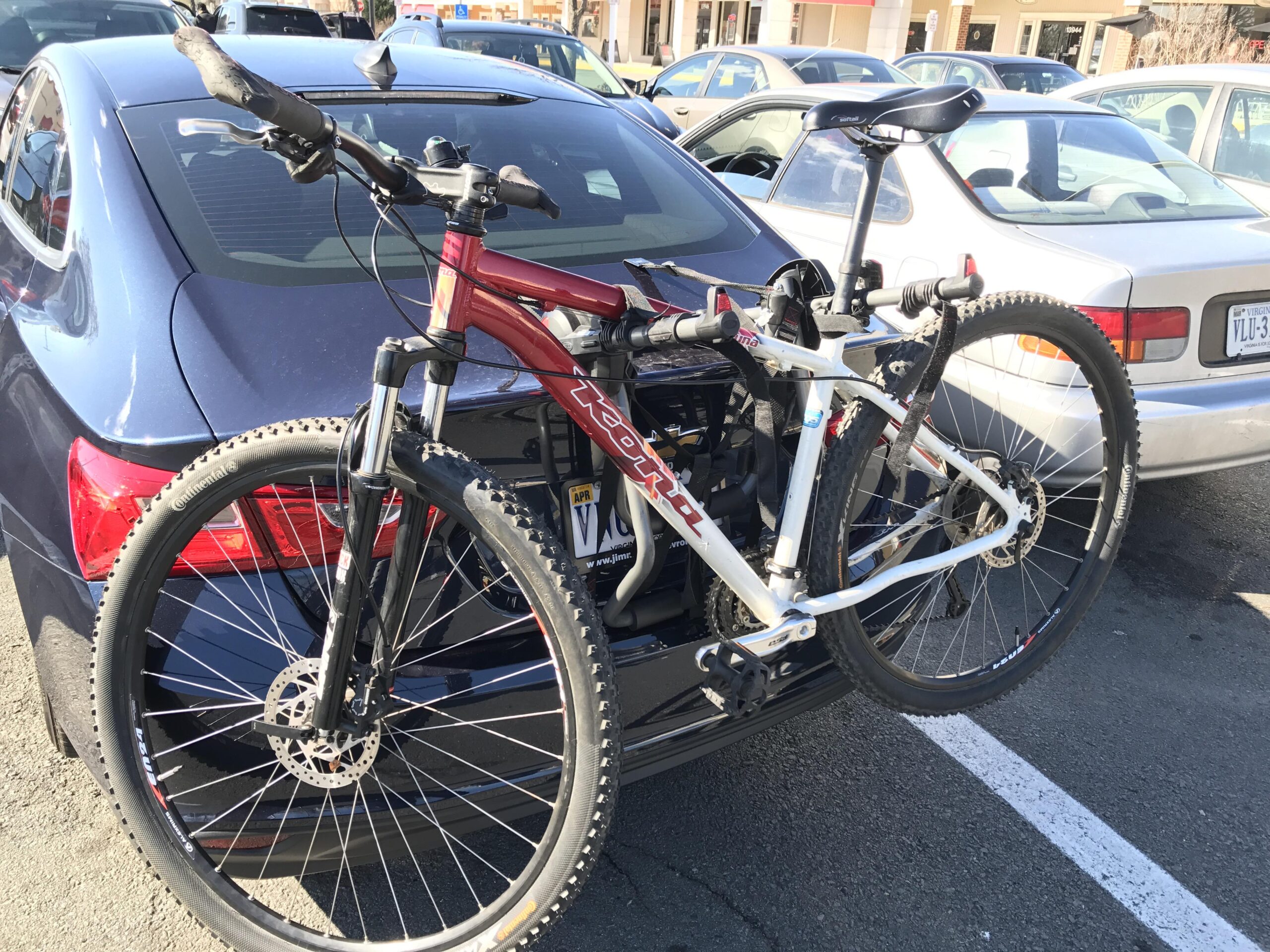 Kona Kahuna: A red and white mountain bike secured on the back of a blue car, parked in a lot. The bike is attached to a bike rack, with one side of the vehicle slightly visible in the background.