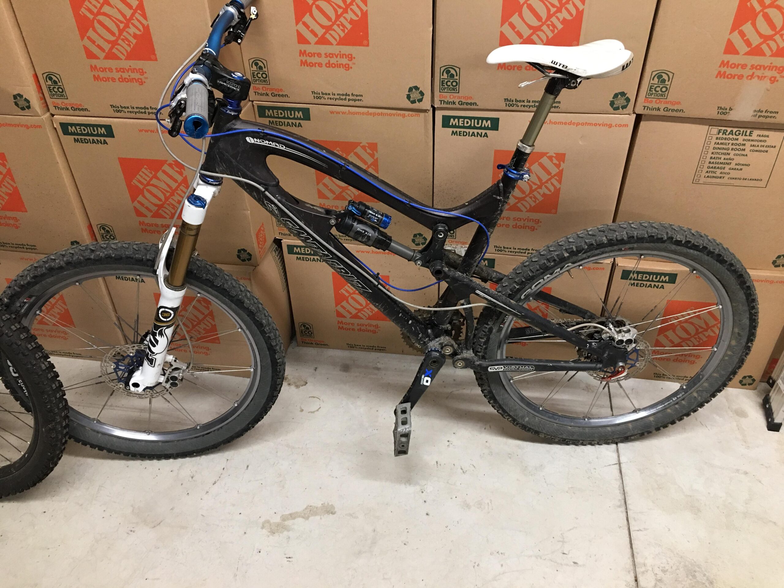 Santa Cruz Nomad: A mountain bike with a black and blue frame, featuring front suspension, disc brakes, and knobby tires, positioned next to cardboard boxes in a storage area. The bike's seat is white, and the handlebars include grips and cables.