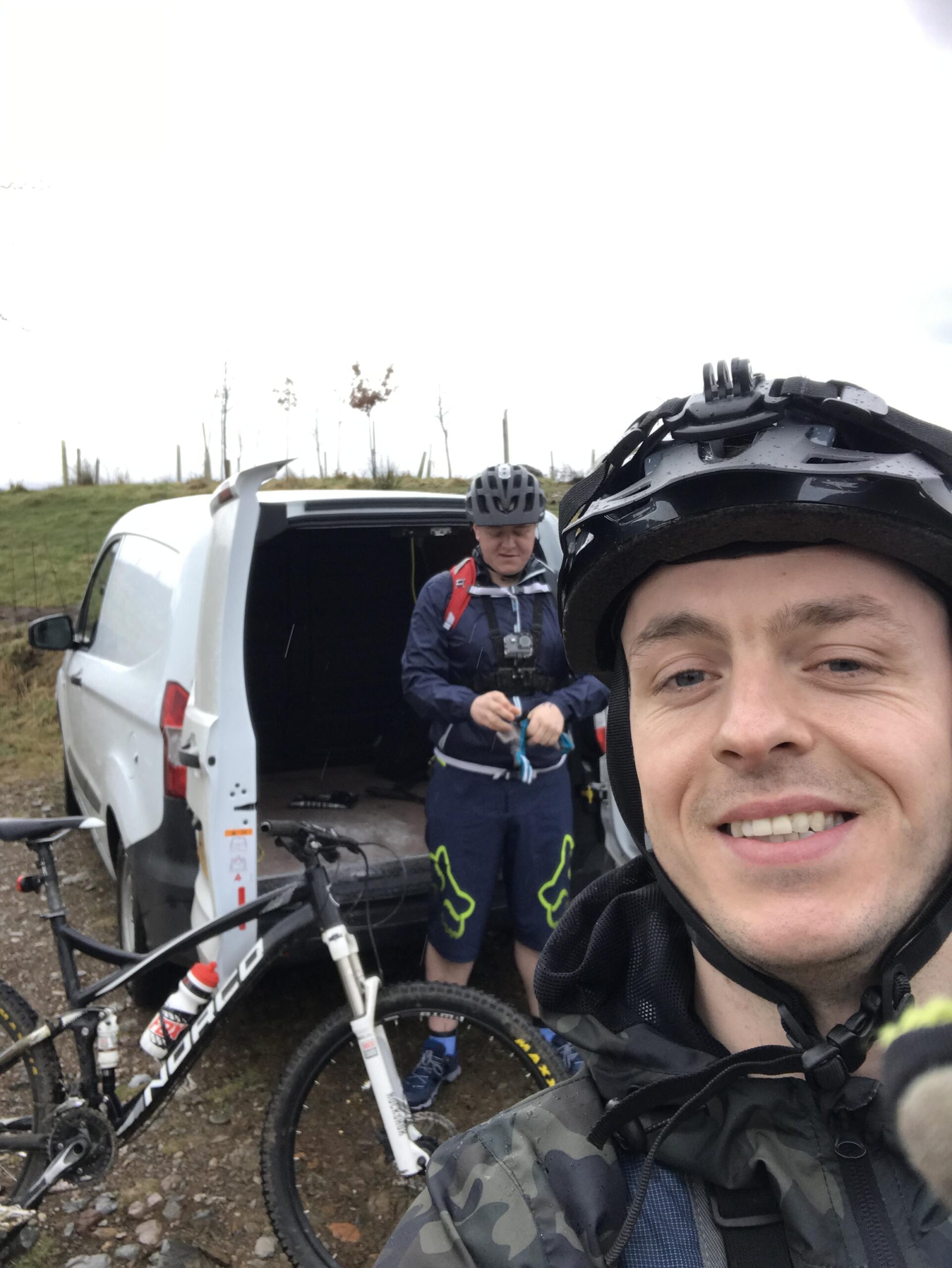 Norco Fluid 7.1: A selfie of two mountain bikers preparing for a ride. In the foreground, a smiling man wearing a black helmet and a camo jacket holds the camera. In the background, another man in a blue jacket and helmet is getting equipment from a white van, with a mountain bike parked beside them. The weather appears overcast and misty, suggesting a cool, damp day.