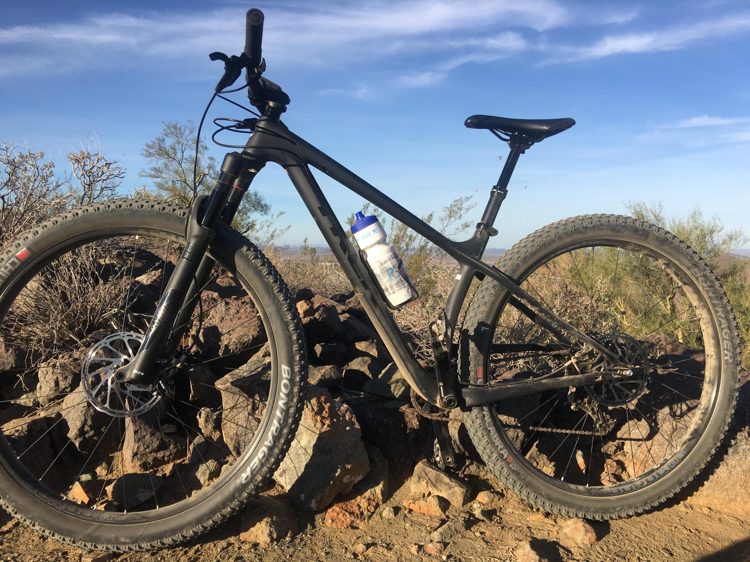 Trek Stache 9 29 Plus: A black mountain bike is leaning against a pile of rocks, with a water bottle attached to its frame. The scene is set outdoors against a backdrop of blue sky and sparse vegetation, suggesting a rugged terrain. The bike has thick tires suitable for off-road riding.