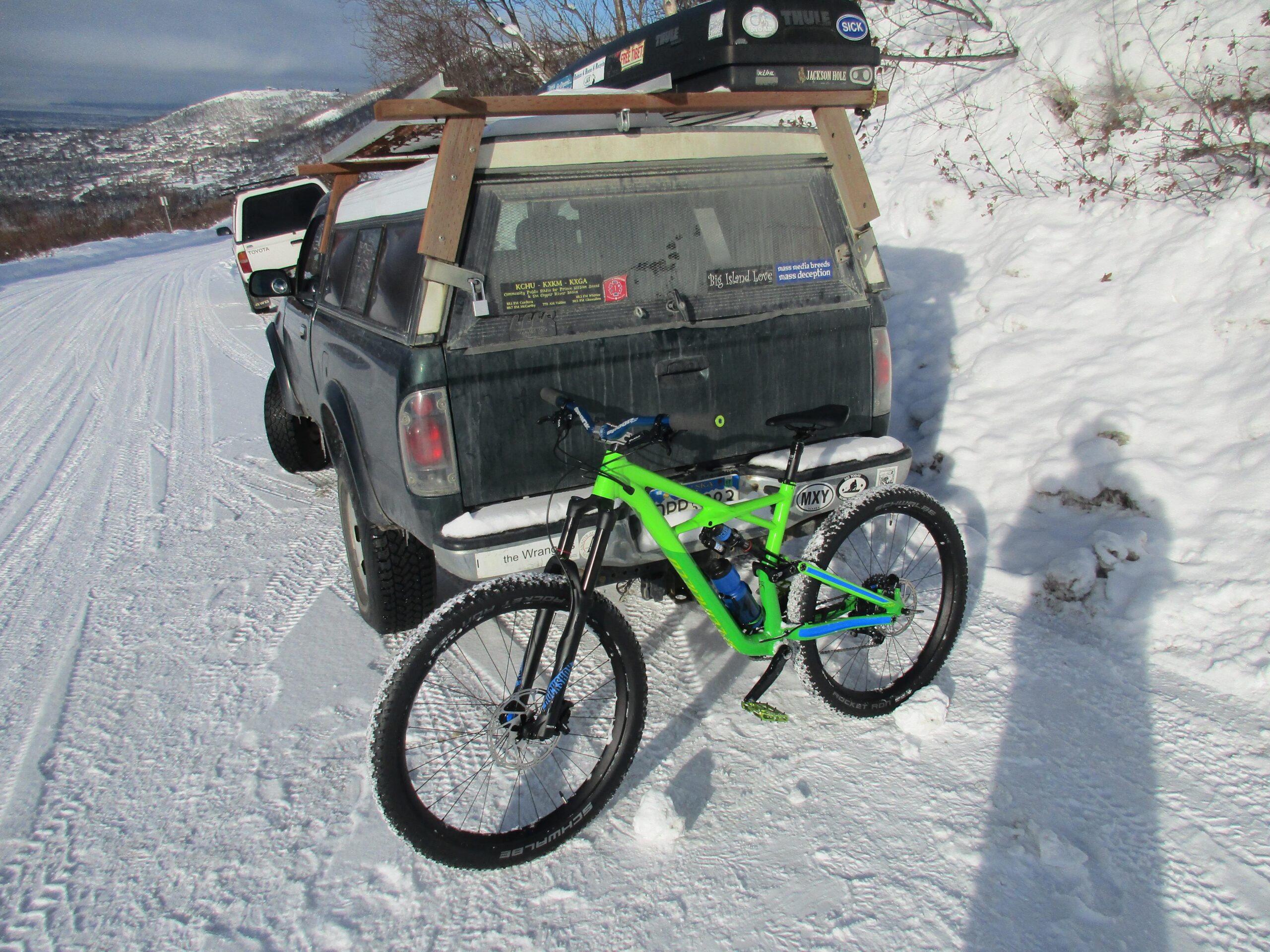 Specialized Enduro Comp 29: A green mountain bike is positioned next to a dirty, parked pickup truck on a snowy road, with snow-covered hills in the background. The truck's back window displays various stickers, and the scene is brightened by natural sunlight.