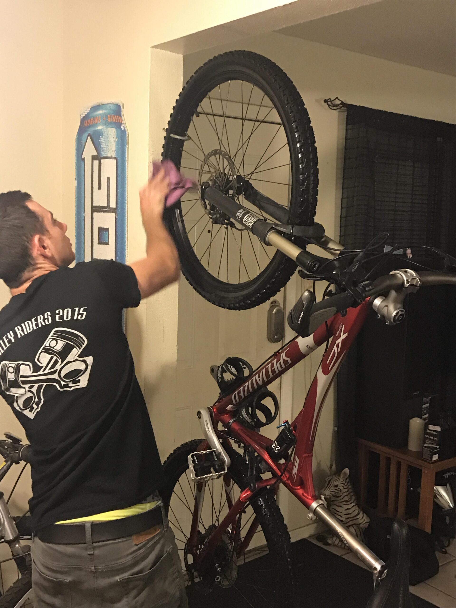 Specialized FSR XC Comp: A person wearing a black t-shirt is cleaning the wheel of a mountain bike hanging on a wall. The bike has a red frame with black wheels and is positioned next to a door. In the background, there is a wall with a painted can design and a faint outline of a small white decorative object resembling a tiger.