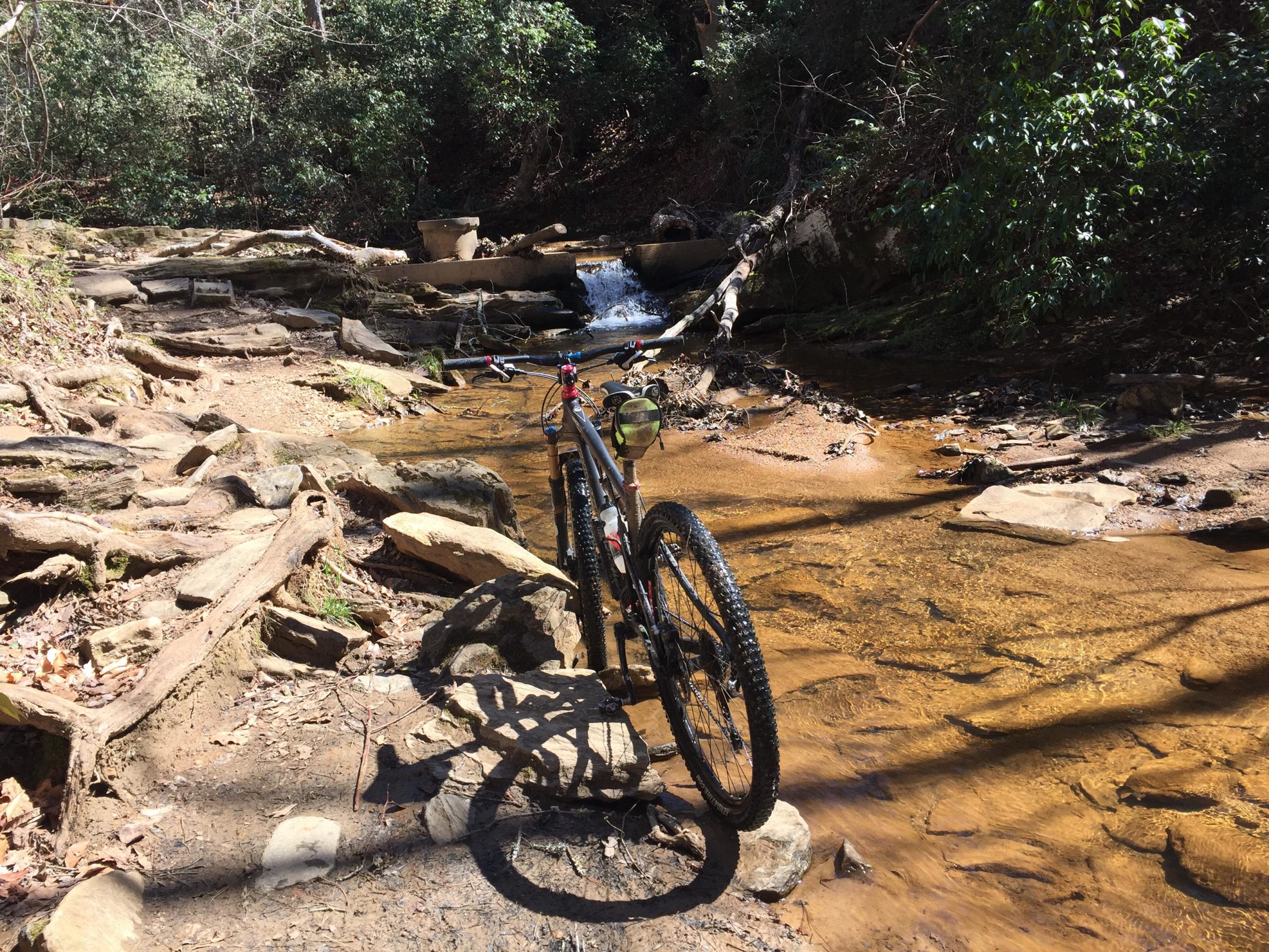 A mountain bike parked near a shallow creek, surrounded by rocky terrain and lush greenery. The sun shines down, creating a warm atmosphere, while a small waterfall cascades in the background. Chicopee Woods mountain bike trail.