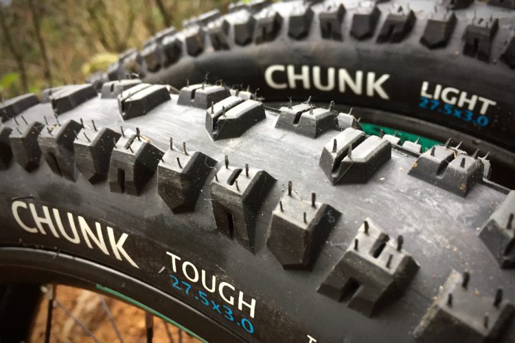 Close-up of two mountain bike tires displaying their tread patterns and branding. One tire is labeled "CHUNK TOUGH" and the other "CHUNK LIGHT," both featuring prominent, angular treads designed for traction. The size of the tires is noted as 27.5 x 3.0, indicating they are suitable for rugged terrain. The background shows blurred greenery, suggesting an outdoor setting.