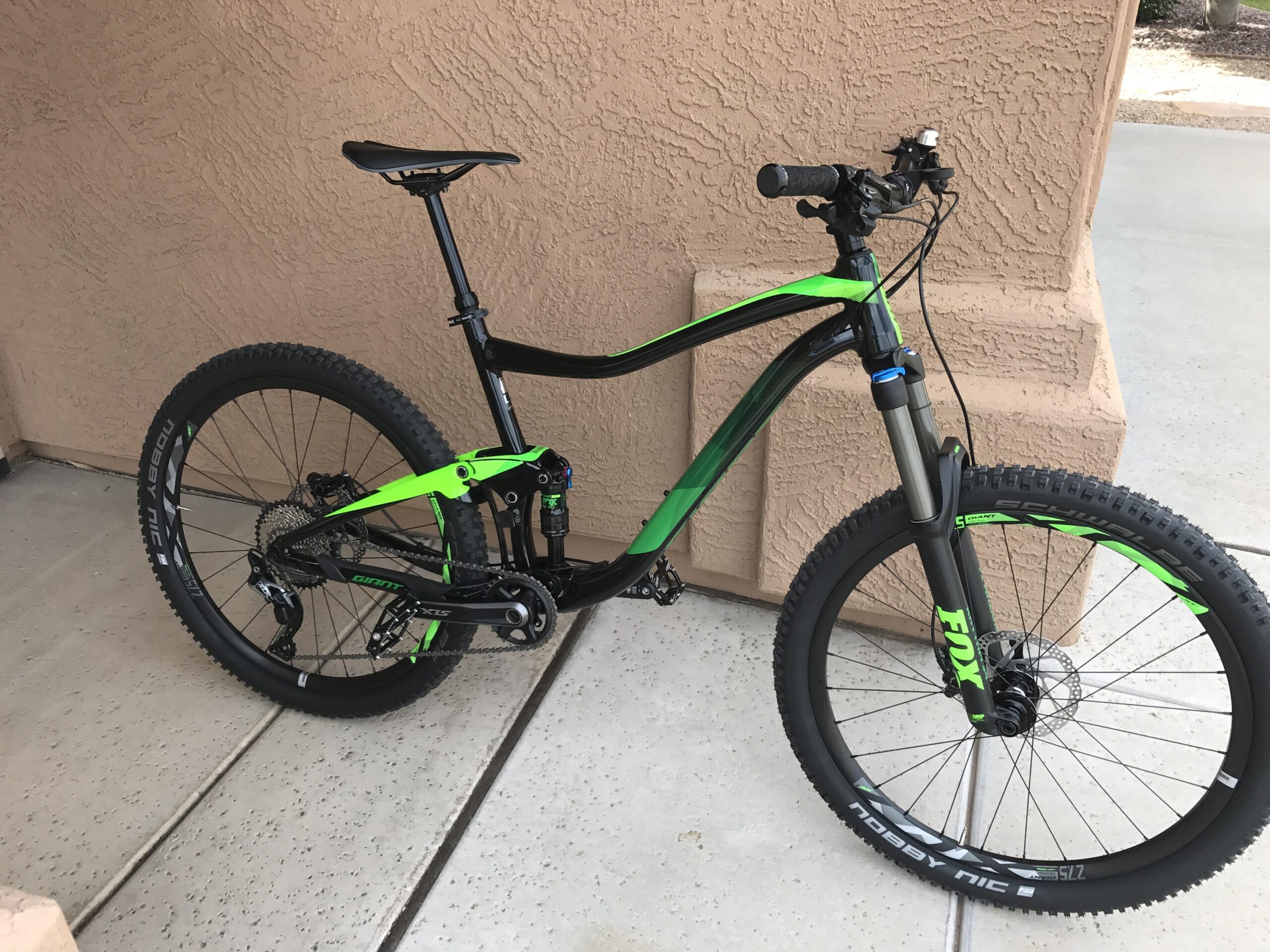 Giant Trance 27.5 2: Mountain bike with a sleek black and green design, featuring a high-performance frame, front suspension, disc brakes, and rugged tires. The bike is positioned on a concrete surface near a textured wall, highlighting its sporty features and modern style.