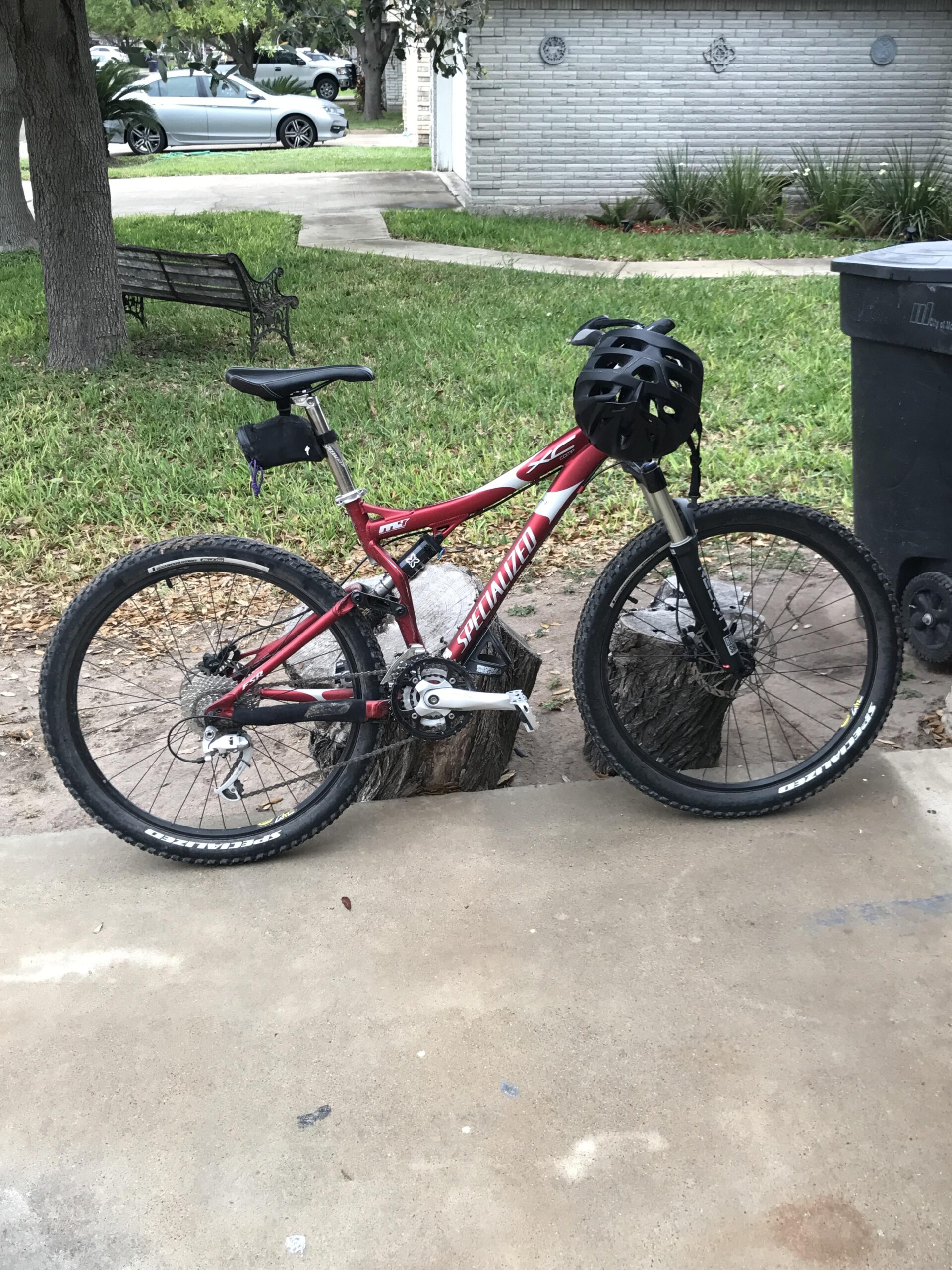 Specialized FSR XC Comp: A red mountain bike with a black helmet attached to the handlebars, resting on a concrete surface next to a tree stump. In the background, there is a grassy area, a parked car, and a bench.