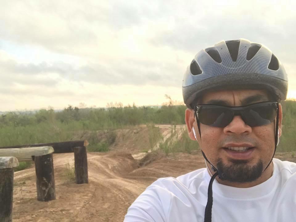 A man wearing a helmet and sunglasses takes a selfie on a dirt biking trail, with a landscape of greenery and cloudy skies in the background. Mission Trails mountain bike trail.