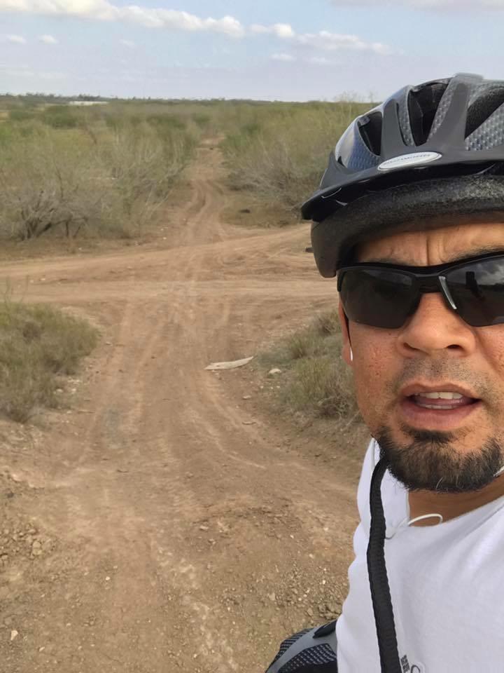 A cyclist wearing a helmet and sunglasses stands on a dirt path with an outdoor landscape in the background. The path splits into two directions, surrounded by greenery and open fields under a partly cloudy sky. Mission Trails mountain bike trail.