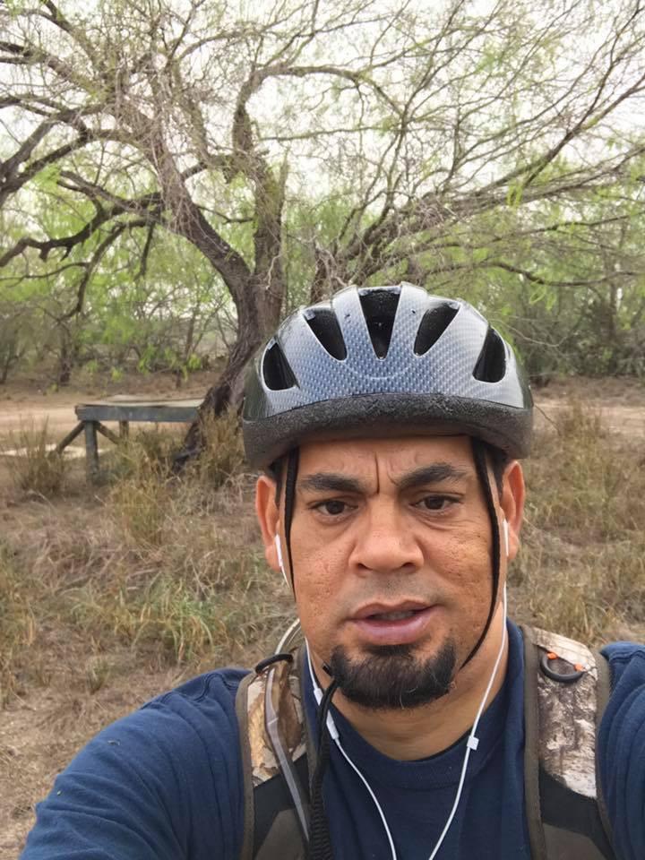 A person wearing a bicycle helmet and earphones is taking a selfie outdoors, surrounded by trees and tall grass. In the background, there is a wooden bench or platform partially visible. The setting appears to be natural and serene. Mission Trails mountain bike trail.