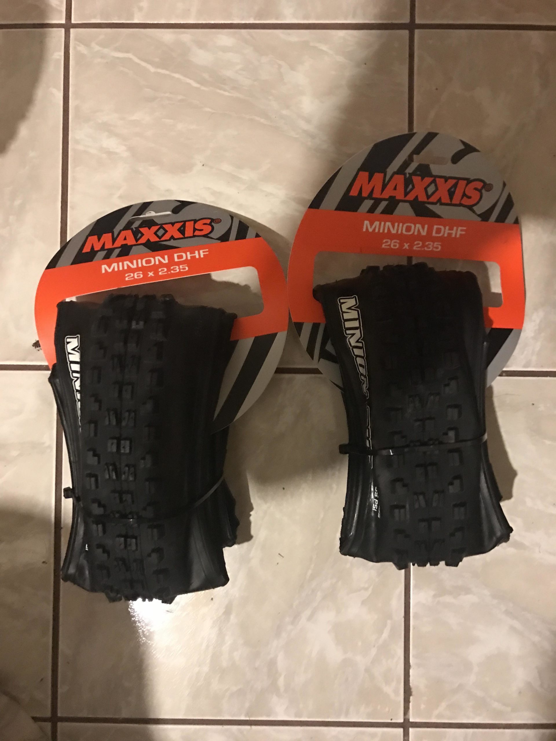 Two Maxxis Minion DHF mountain bike tires, each measuring 26 x 2.35, are displayed on a tiled floor. The tires are mounted on circular cardboard packaging featuring the Maxxis logo and product information. Mission Trails mountain bike trail.