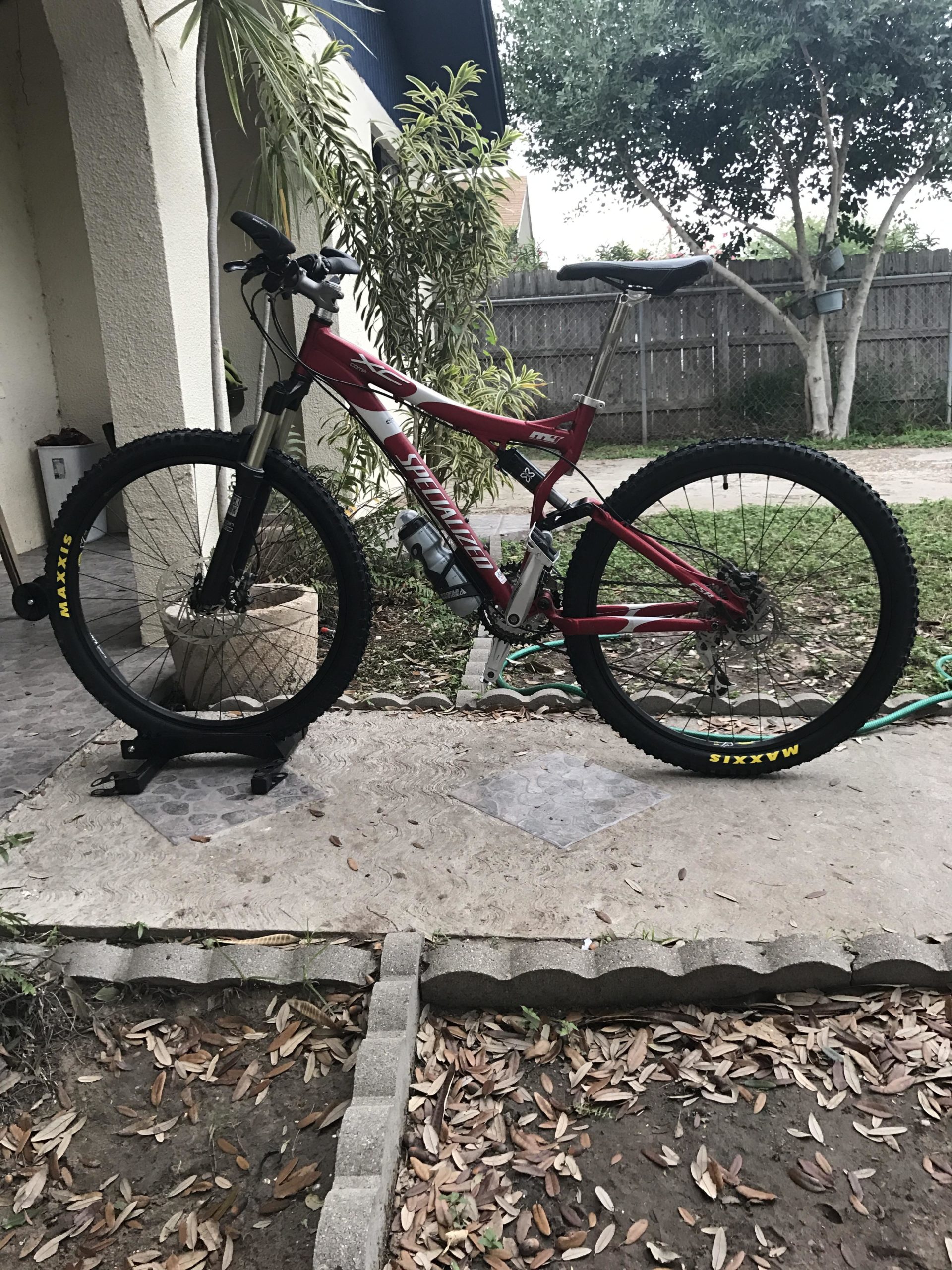 A red and white mountain bike with wide tires is parked on a stone surface, next to a house. There are plants in the background and a small garden area with fallen leaves. The handlebars are turned to the side, and the bike is positioned on a stand. Mission Trails mountain bike trail.
