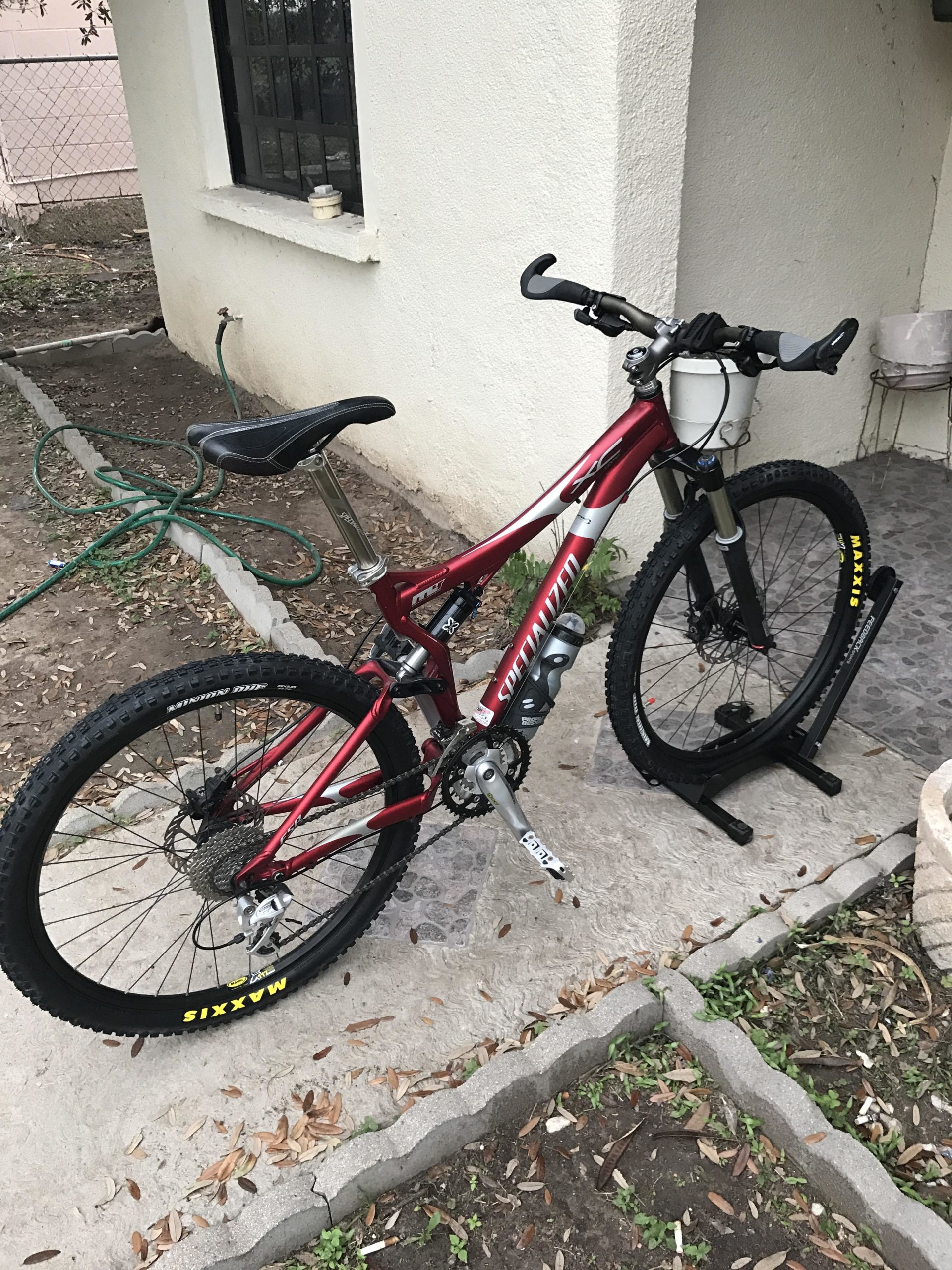 A red mountain bike parked next to a beige wall with a window. The bike features wide tires with yellow markings, a prominent seat, and handlebars. There is a garden hose on the ground, and the surrounding area has a mix of concrete and dirt, with some fallen leaves visible. Mission Trails mountain bike trail.