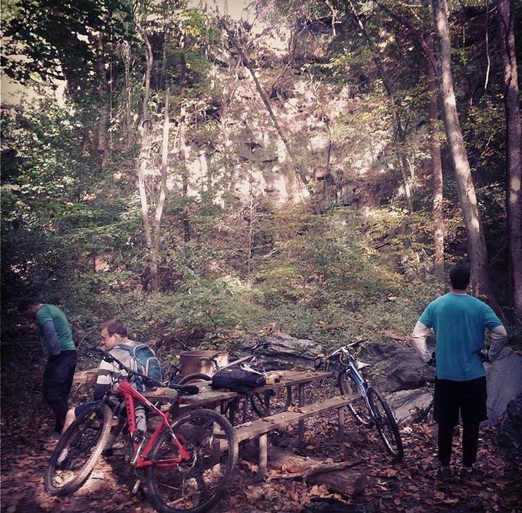 A group of three cyclists stands in a wooded area, surrounded by trees and rocks. Two bikes are resting on a wooden bench near them, while the cyclists appear engaged in conversation or observation of the landscape. The scene is set in a natural environment with fallen leaves on the ground and a steep rock face in the background. Smedley park mountain bike trail.