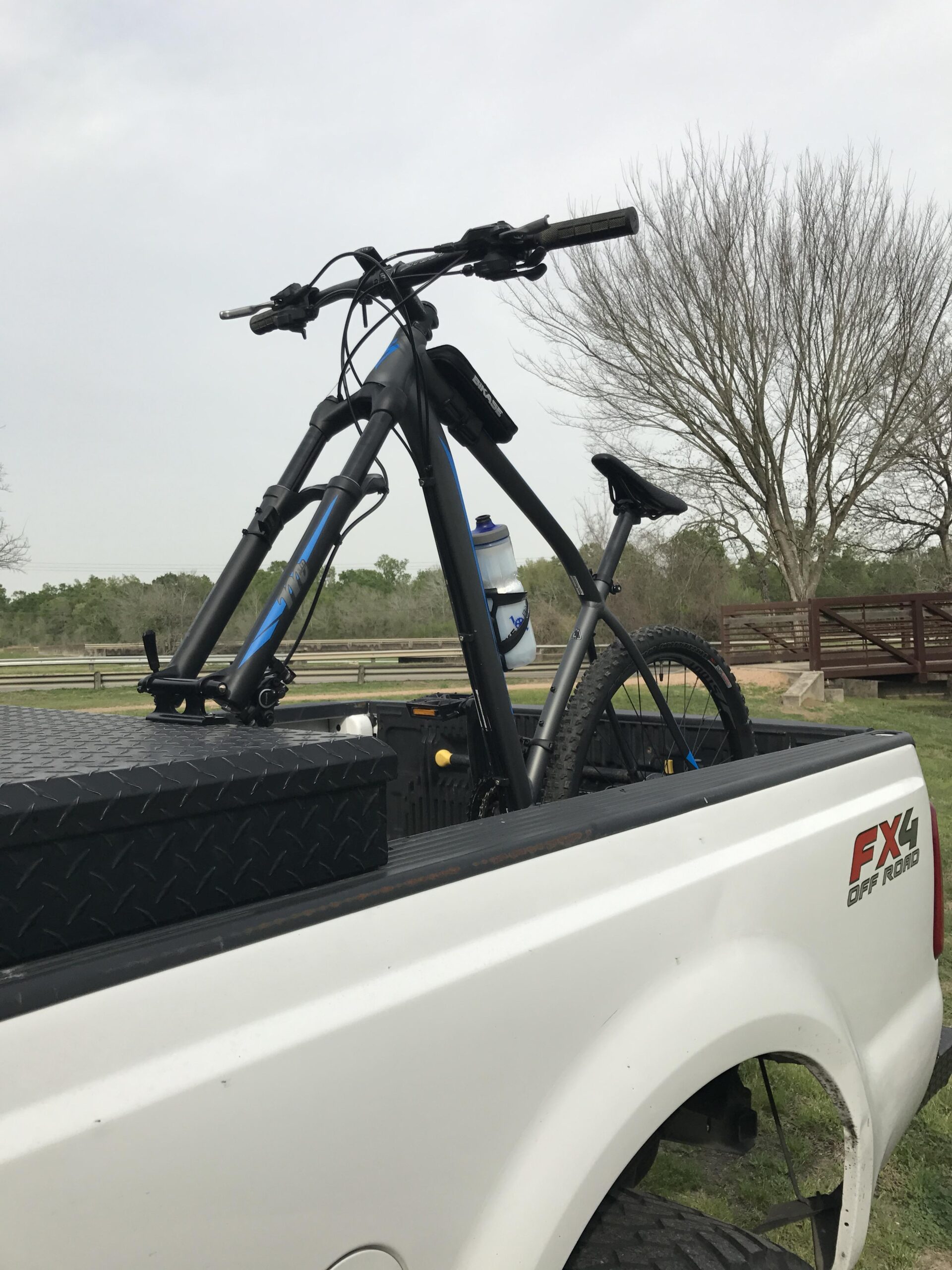 Specialized Rockhopper Expert Disc 29er: A black mountain bike with blue accents is secured in the bed of a white pickup truck. A water bottle is attached to the bike, and a textured black storage box is also visible in the truck bed. The background features a sparse landscape with a leafless tree and a cloudy sky.