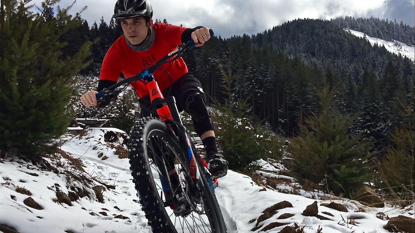 Maxxis Forekaster: A cyclist in a red shirt and black gear rides a mountain bike along a snowy trail surrounded by greenery and mountains under a partly cloudy sky.