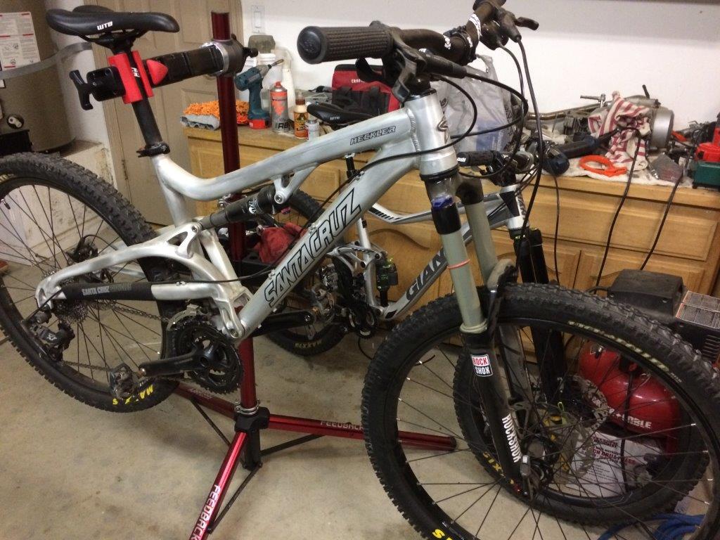 Santa Cruz Heckler: A close-up view of a silver mountain bike, specifically a Santa Cruz model, positioned on a repair stand in a workshop. The bike features prominent components like a suspension fork and thick tires. The background includes various tools and equipment typical of a bike maintenance area.