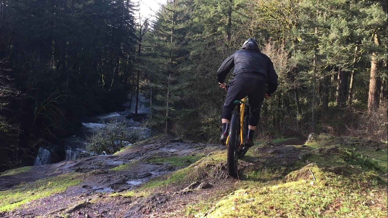 A mountain biker riding through a forested trail, navigating a rocky area near a flowing stream. The scene features lush greenery and tall trees, capturing the essence of outdoor adventure. Round Lake mountain bike trail.
