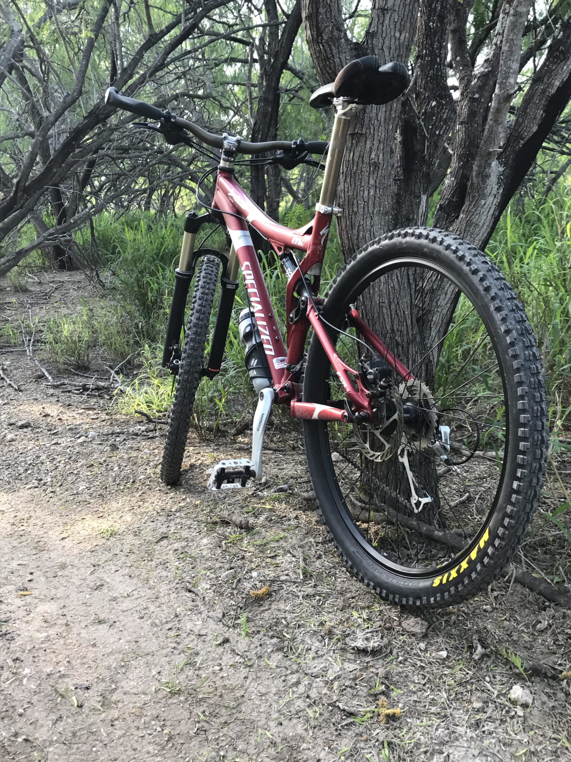 A red mountain bike is leaning against a tree in a wooded area, surrounded by green grass and bushes. The bike features a sturdy frame, thick tires, and is positioned on a dirt path. Nearby branches and foliage create a natural outdoor setting. Mission Trails mountain bike trail.