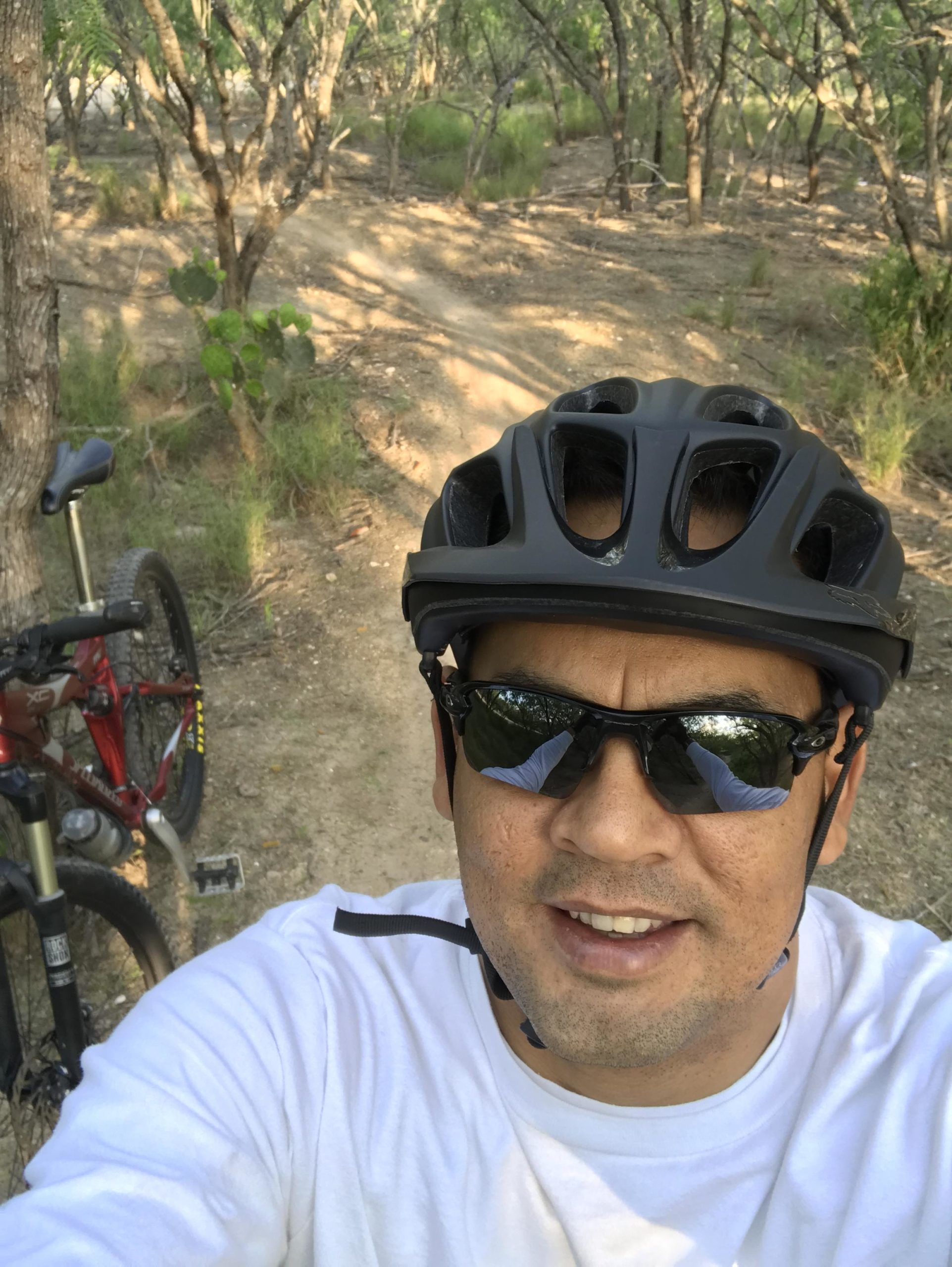 A person wearing a black bicycle helmet and sunglasses smiles for a selfie. They are standing on a dirt path surrounded by trees and vegetation, with a red mountain bike parked beside them. The setting appears to be a natural outdoor environment, ideal for biking. Mission Trails mountain bike trail.