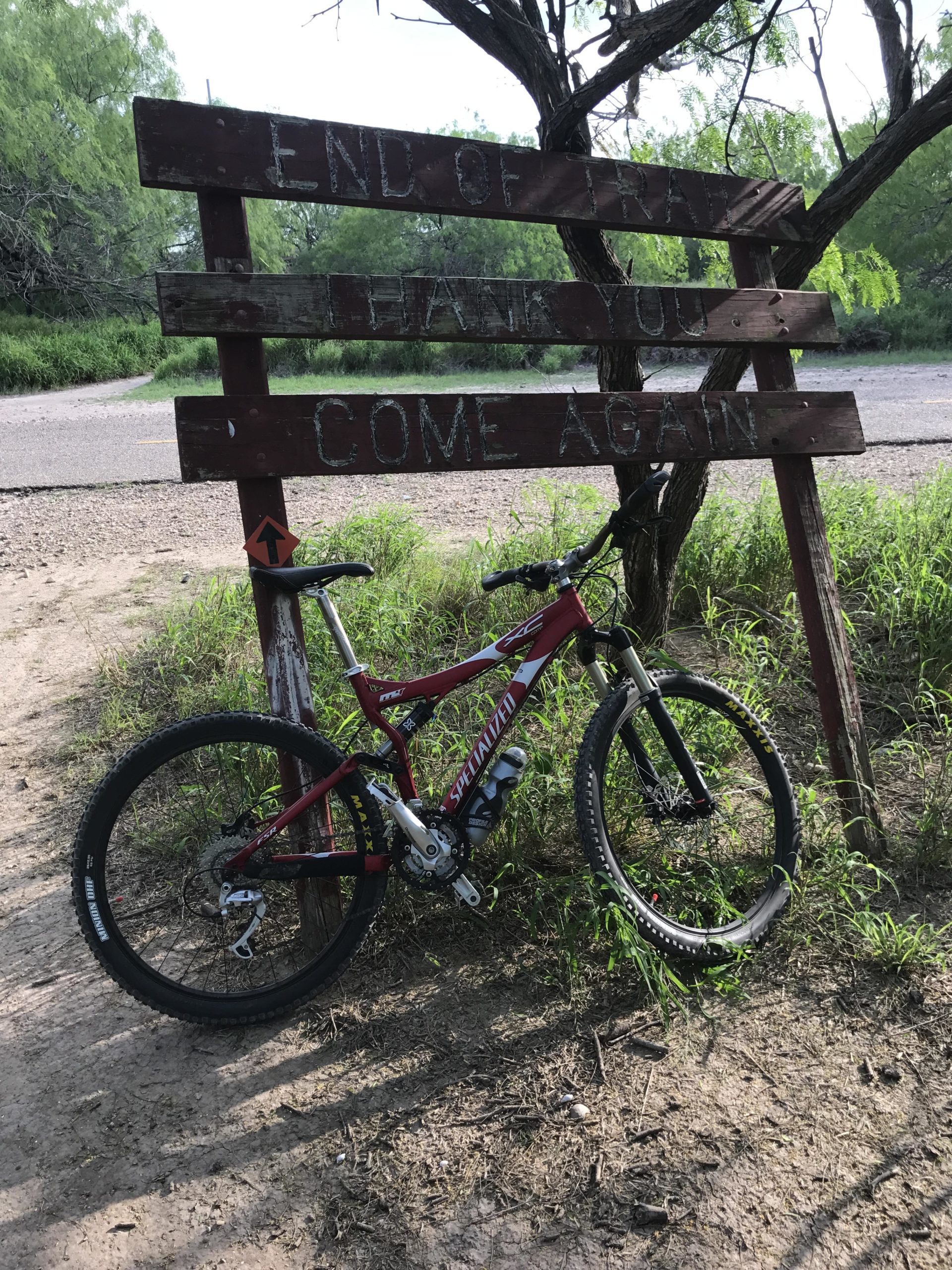 A mountain bike rests against a wooden sign that reads "END OF TRAIL" at a nature path's conclusion, surrounded by lush greenery and a dirt trail. The sign also says "THANK YOU" and "COME AGAIN," indicating the end of the biking route. Mission Trails mountain bike trail.
