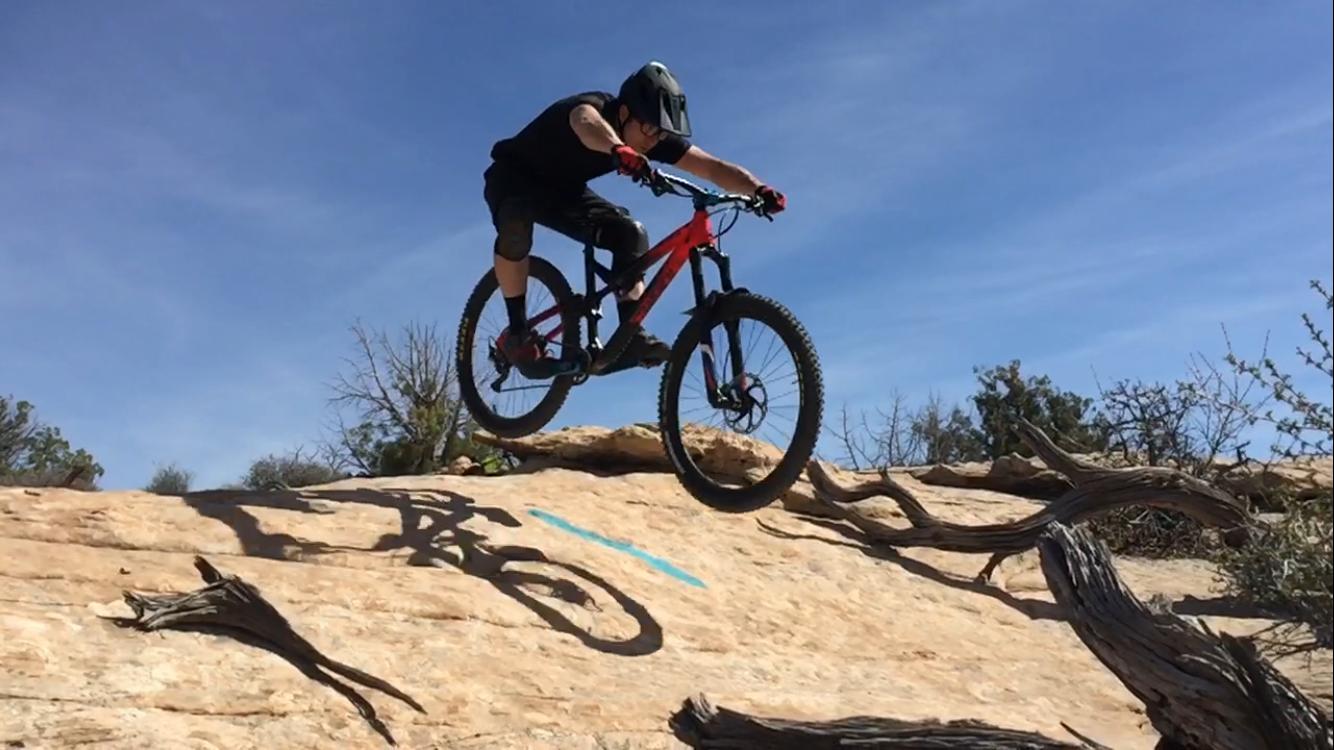 A mountain biker mid-air, performing a jump on rocky terrain. The biker is wearing a helmet and protective gear, with a black and red mountain bike. The background features a clear blue sky and sparse desert vegetation. Navajo Rocks mountain bike trail.