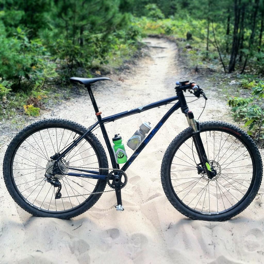 A mountain bike with a black frame is parked on a sandy trail surrounded by greenery. Two water bottles are mounted on the bike, one green and one gray. The background features a winding dirt path leading into the woods.