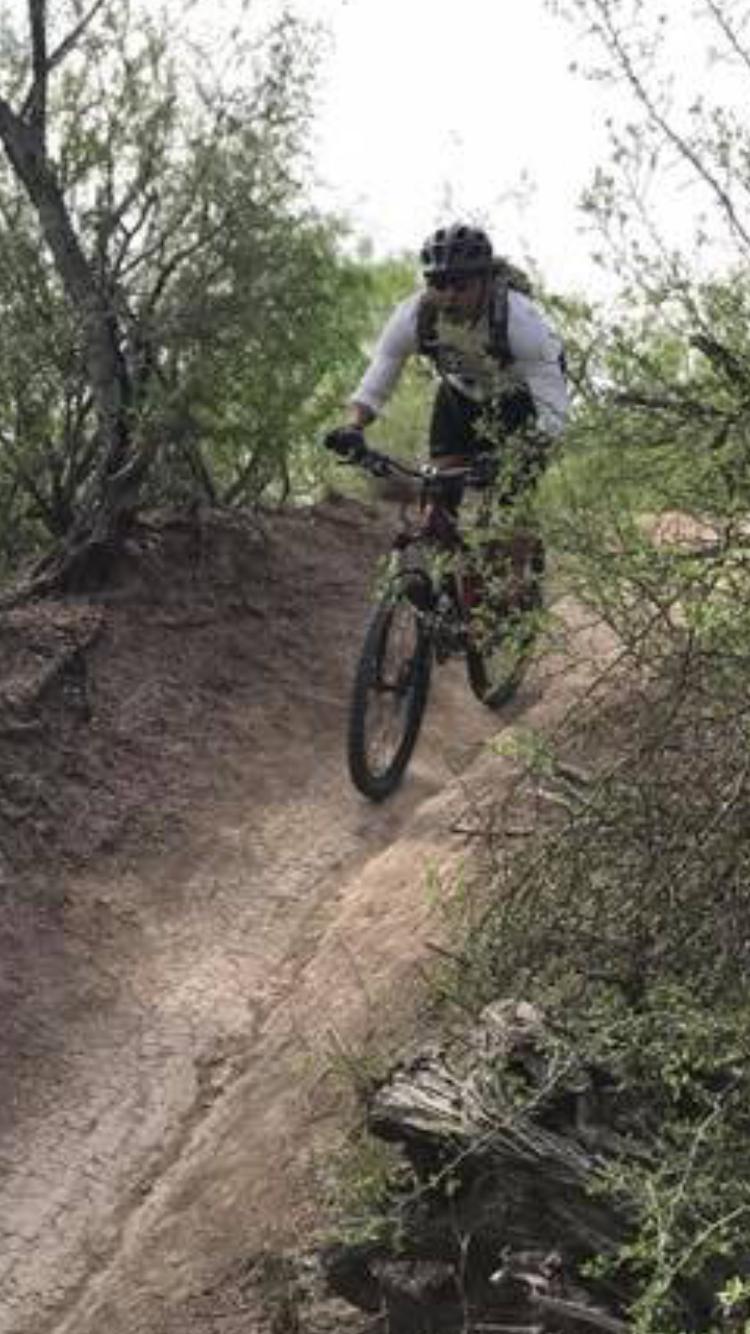 A person riding a mountain bike down a dirt trail surrounded by shrubs and trees. The rider is wearing a helmet and protective gear, navigating over a slight incline on a rugged, natural path. Mission Trails mountain bike trail.