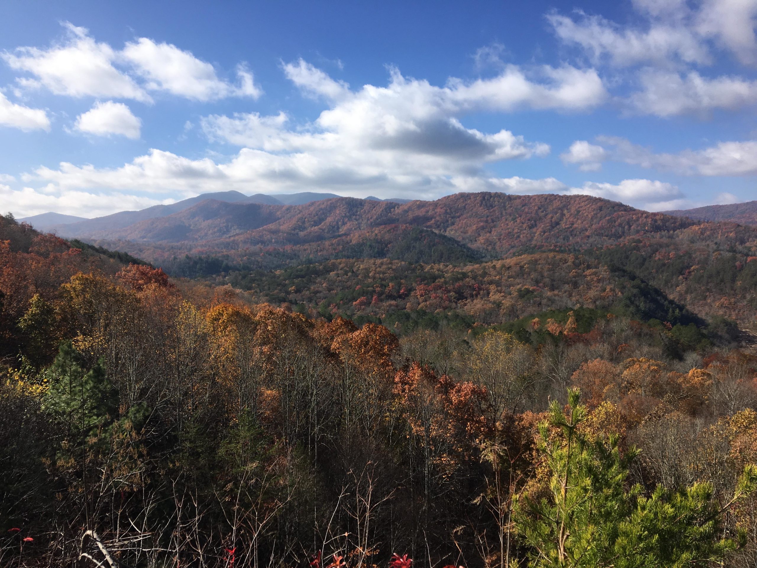 A scenic view of mountains blanketed in autumn foliage, showcasing vibrant reds, oranges, and yellows. The sky is partly cloudy, with sunlight illuminating the landscape, while the rolling hills create a serene backdrop. Brush Creek mountain bike trail.