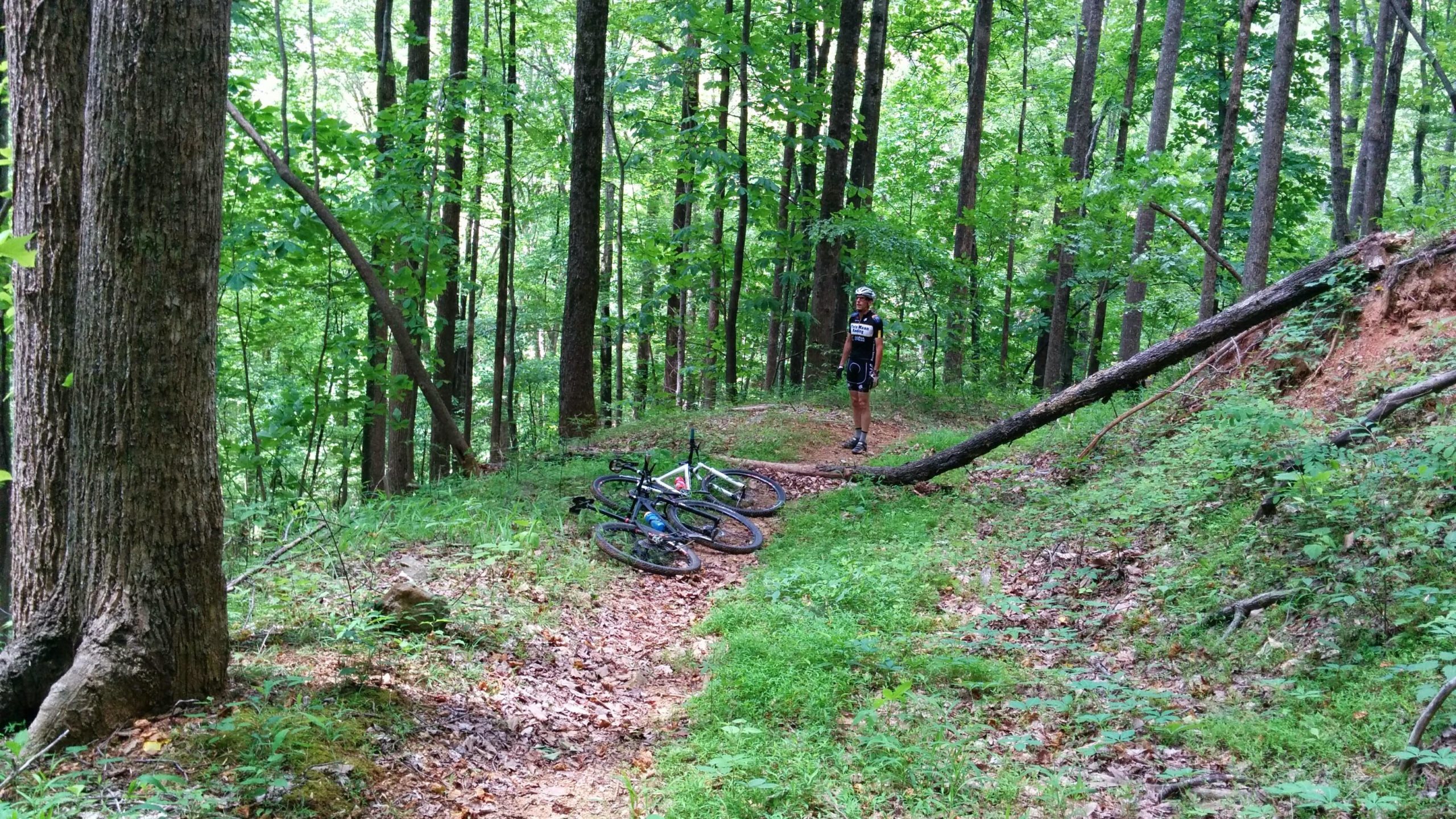 A cyclist in a black and white outfit stands on a dirt path surrounded by tall green trees in a forest. Two mountain bikes are resting on the ground nearby, with sunlight filtering through the leaves, casting a dappled light on the scene. The area is lush with grass and fallen leaves, creating a tranquil outdoor atmosphere. Gem Mine mountain bike trail.