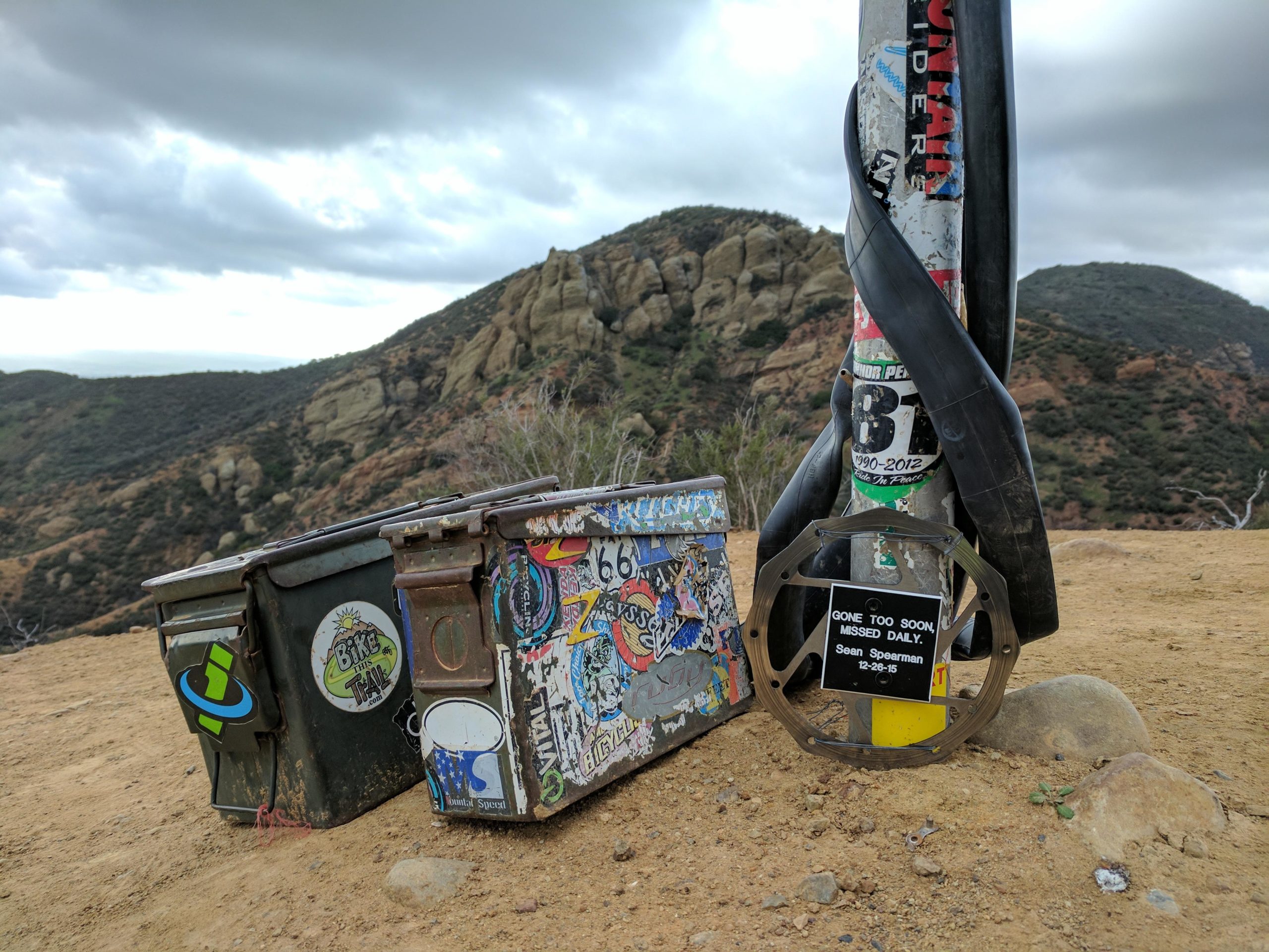 A scenic outdoor view featuring two sticker-covered containers on a dirt path, with mountains in the background under a cloudy sky. One container is green with a "Bike the Trail" sticker, and the other is blue adorned with various stickers. Adjacent to the containers stands a pole wrapped in black tape, displaying a commemorative plaque that reads "GONE TOO SOON, MISSED DAILY. Sean Spearman. 12-26-15." The Luge mountain bike trail.