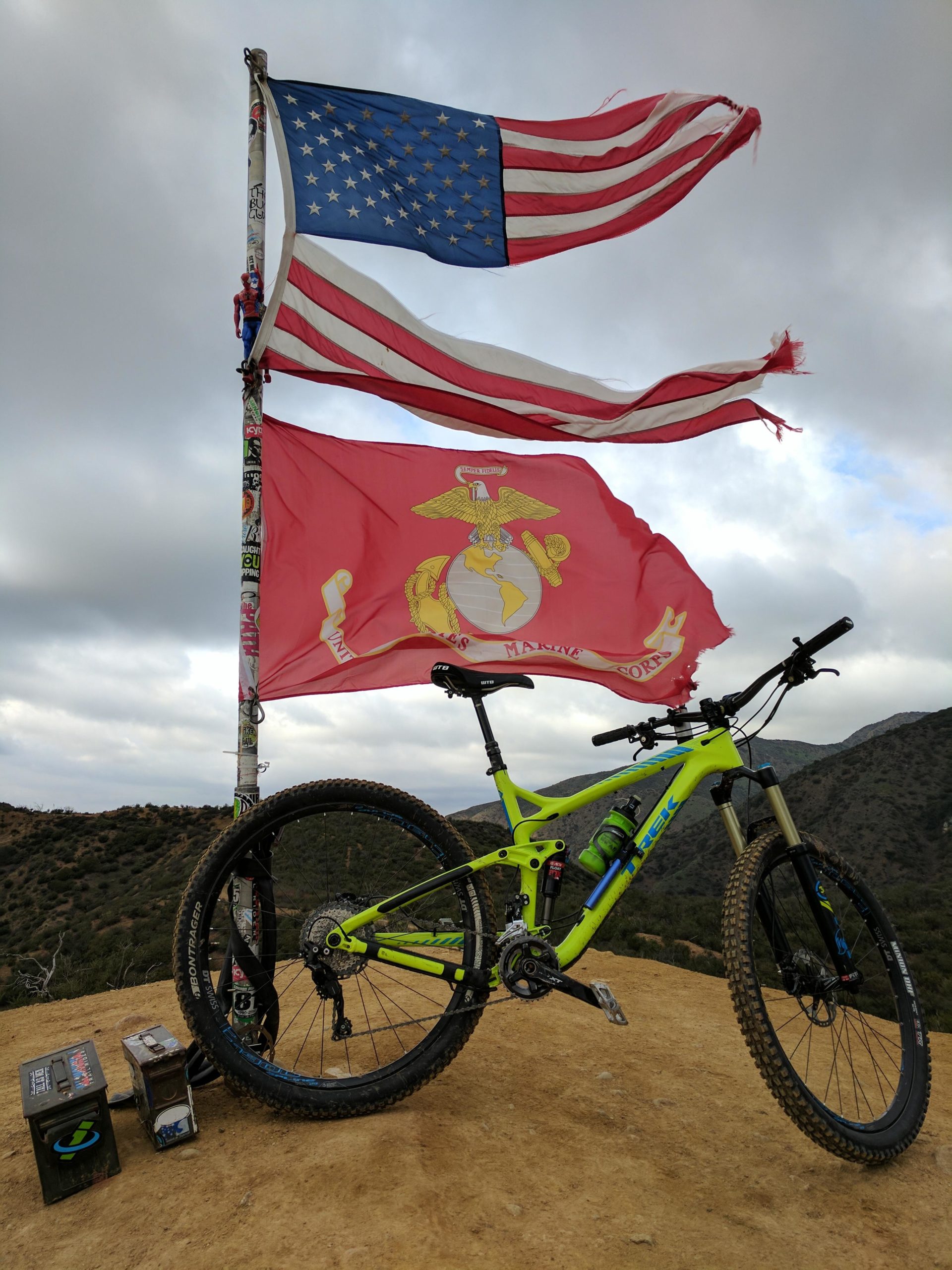 A mountain bike in vivid green color parked on a rocky hillside, with American and Marine Corps flags waving above it. The scene features a cloudy sky and a mountainous landscape in the background, along with two small boxes positioned beside the bike. The Luge mountain bike trail.