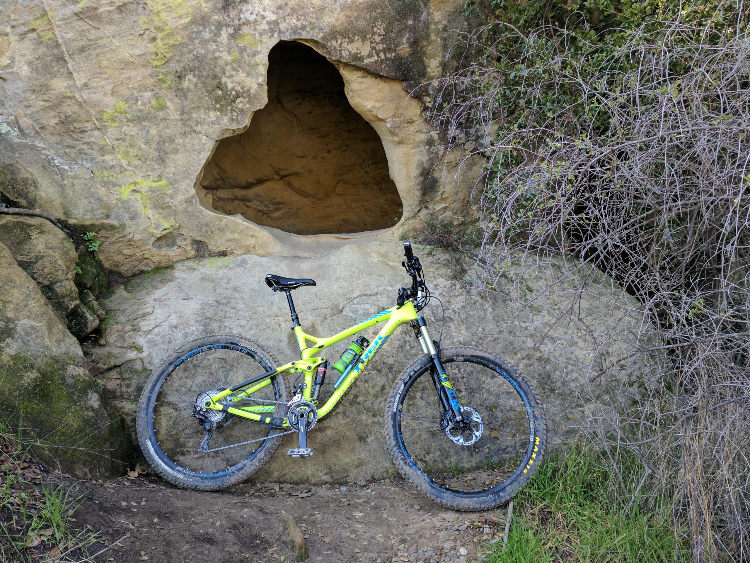 A bright yellow mountain bike rests against a rocky hillside, featuring a natural cave opening in the stone behind it. The surrounding area includes sparse vegetation and dry branches, with hints of greenery on the ground. El Moro / Erection Loop mountain bike trail.
