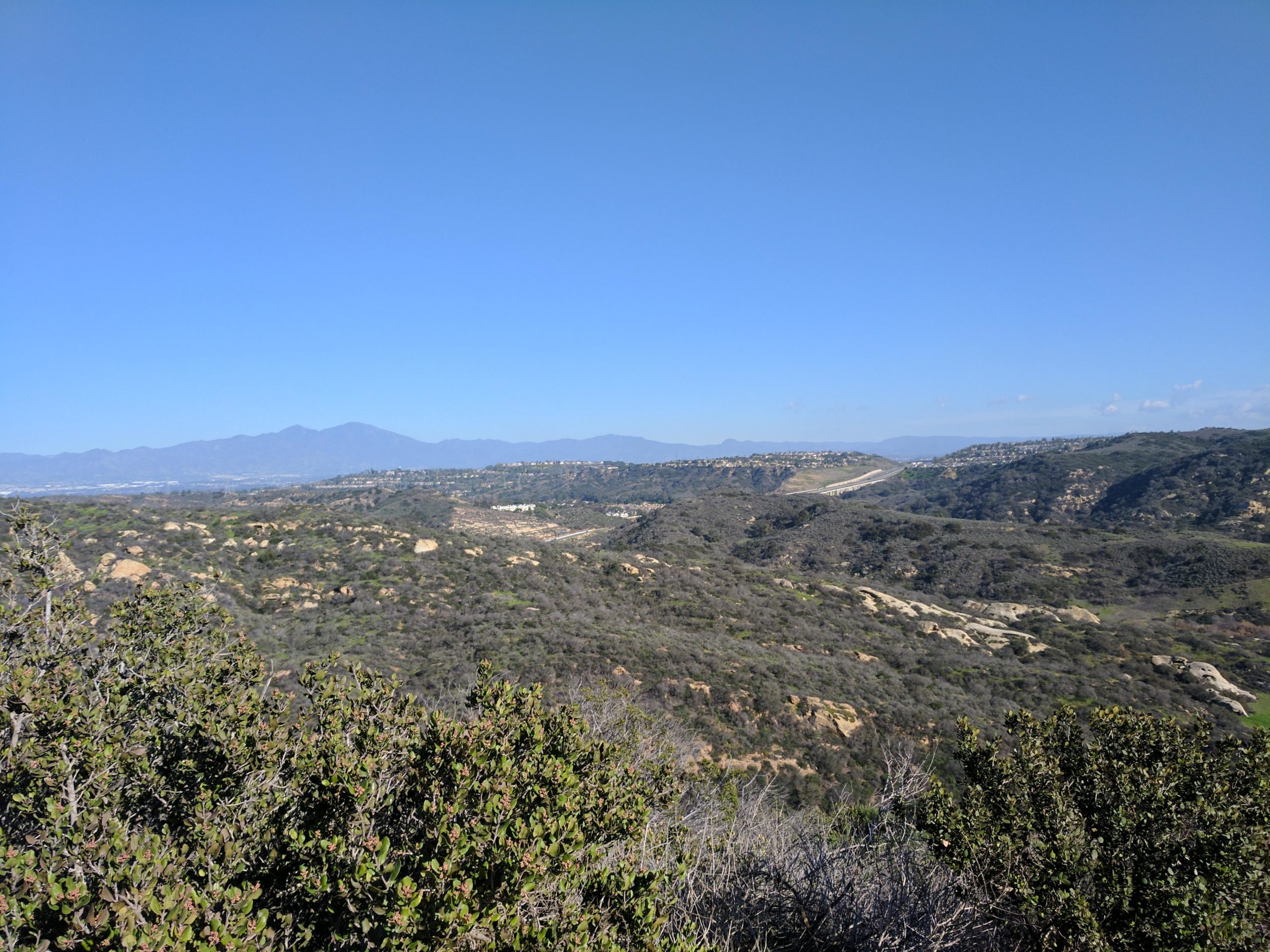 A panoramic view of rolling hills and mountains under a clear blue sky, featuring greenery and rocky outcrops in the foreground, with residential areas visible in the distance along the hillsides. El Moro / Erection Loop mountain bike trail.