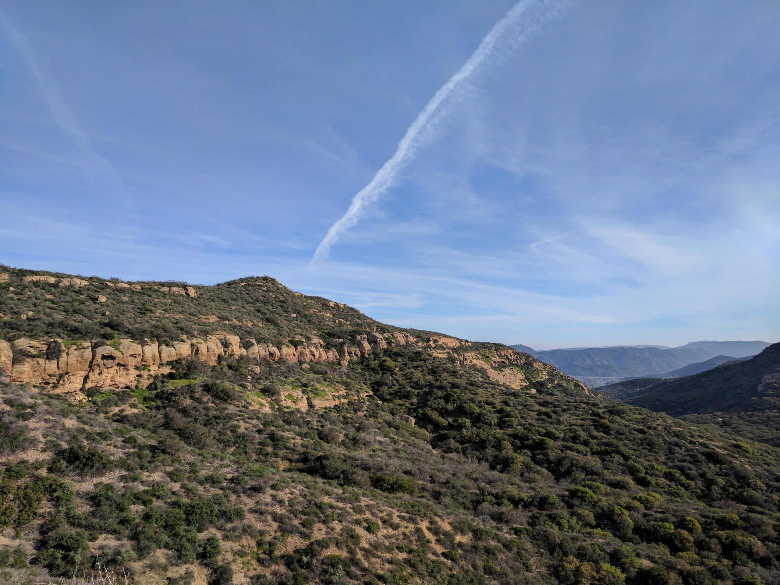 A panoramic view of rolling hills covered with green shrubs and rocky outcrops under a blue sky with wispy clouds. The landscape showcases a natural setting with varying elevations and textures, creating a serene and picturesque environment. El Moro / Erection Loop mountain bike trail.