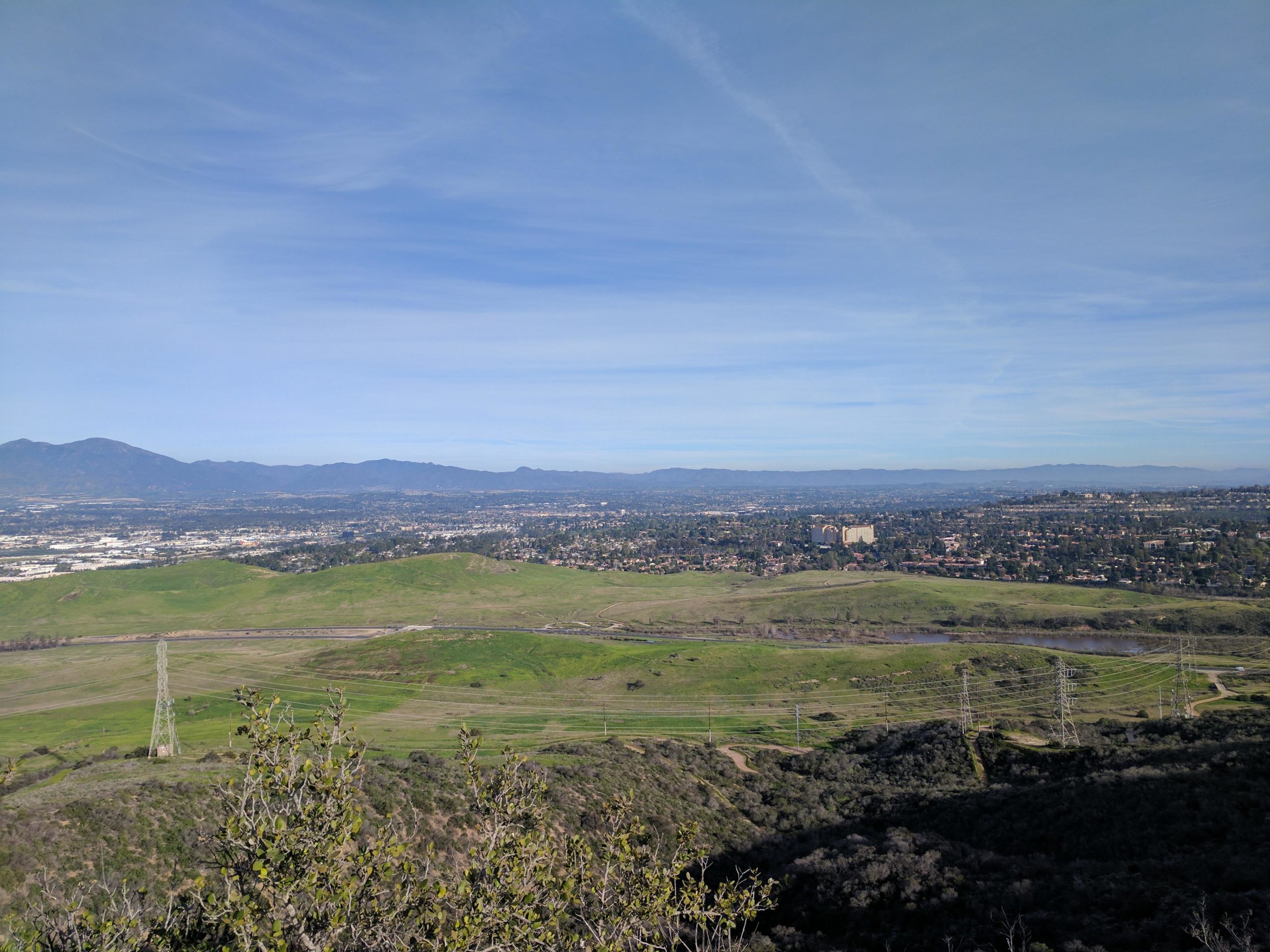 A panoramic view of a green landscape with rolling hills, distant mountains, and a town in the valley below. Power lines are visible in the foreground, and the sky is partly cloudy, with a soft blue hue. El Moro / Erection Loop mountain bike trail.