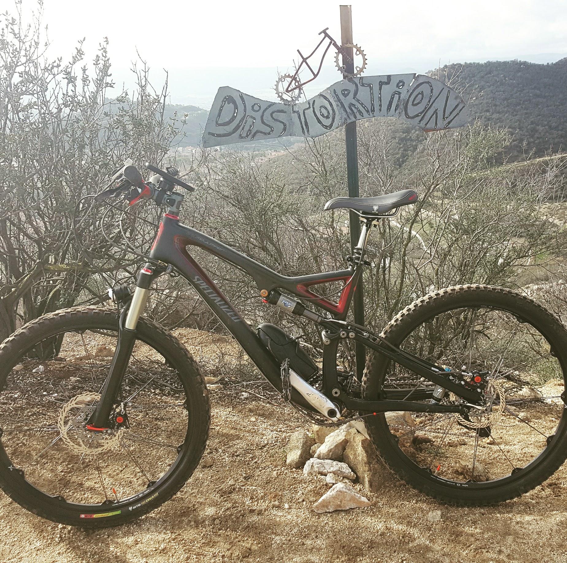 Specialized Stumpjumper FSR Pro: A mountain bike leaning against a sign that reads "Distortion" in a natural outdoor setting, with rocky terrain and sparse vegetation in the background. The scene captures a scenic view of a mountainous landscape under a cloudy sky.