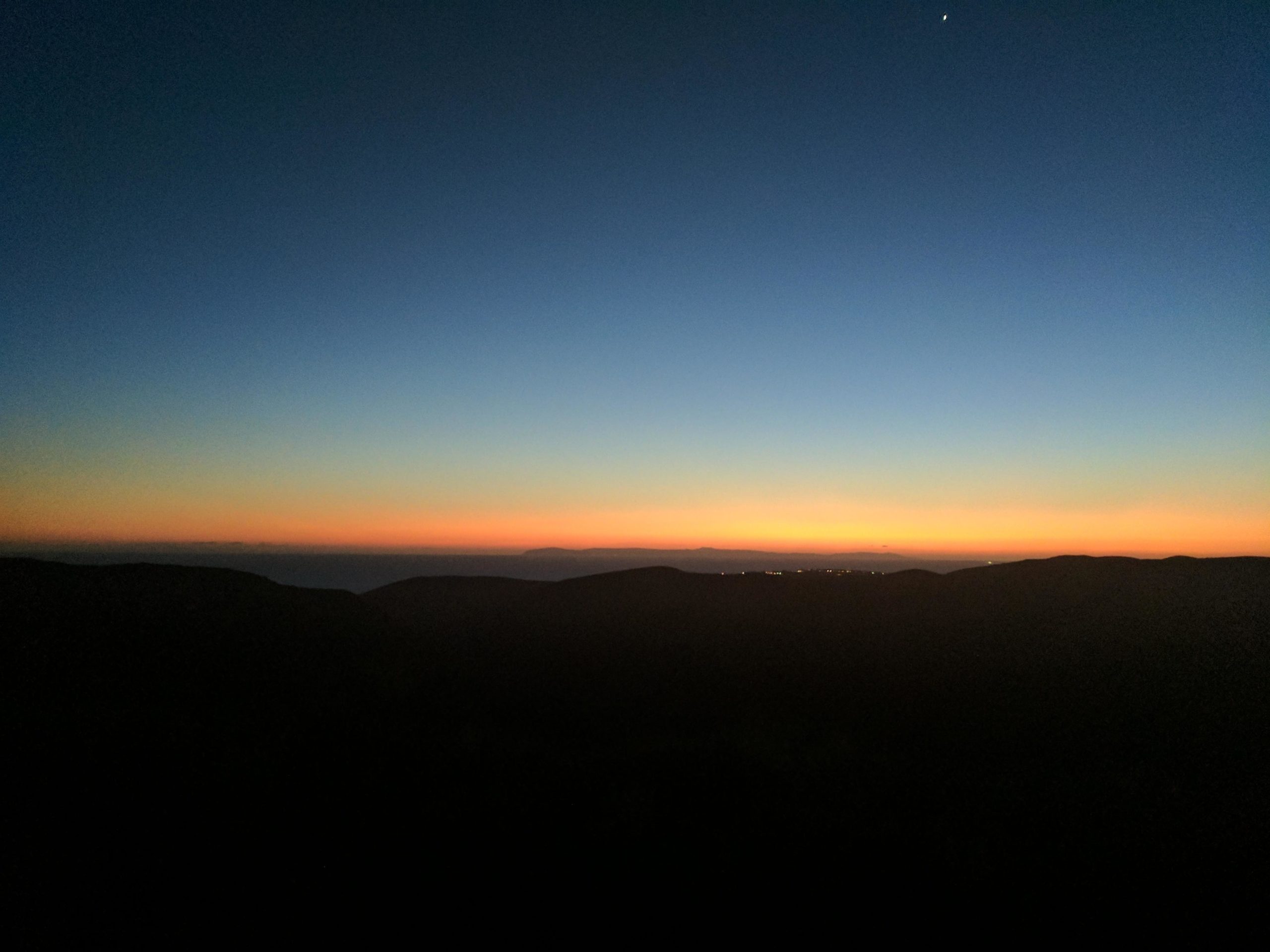 A panoramic view of a twilight sky, transitioning from deep blue at the top to warm orange and yellow hues near the horizon, silhouetted by dark mountain outlines. A small star is visible in the upper right corner. The scene captures the serene beauty of sunset over a landscape. El Moro / Erection Loop mountain bike trail.
