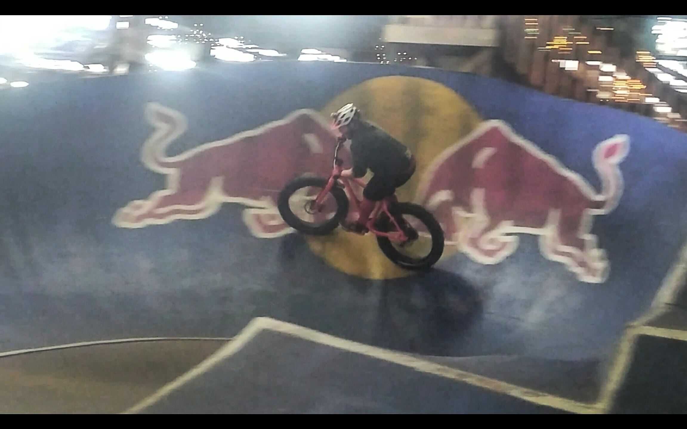 A cyclist performs a trick on a pink fat bike while riding on a curved ramp featuring a Red Bull logo in a dimly lit indoor setting. The rider is wearing a helmet and dark clothing, showcasing a dynamic moment of movement on the ramp. Rays Indoor Mtb Park mountain bike trail.