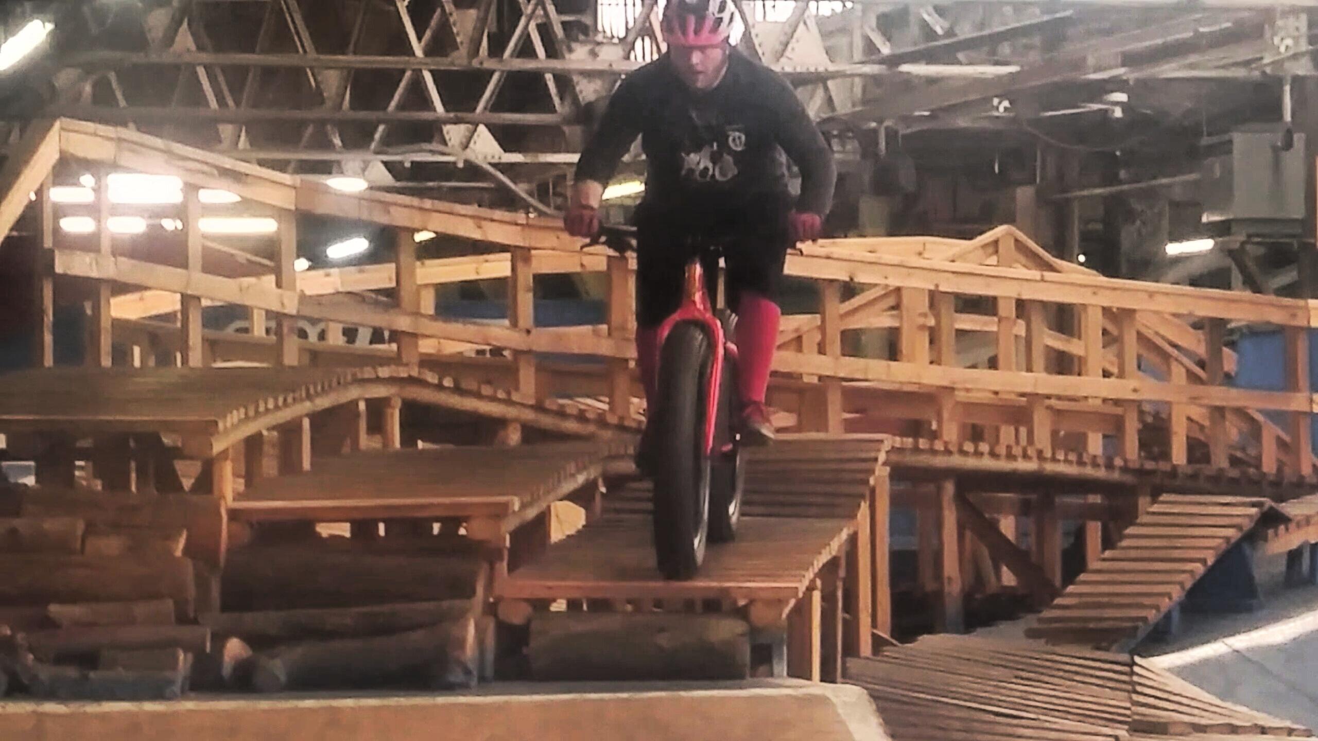 A person riding a fat bike on a wooden obstacle course inside a warehouse. The scene features various ramps and wooden structures, with the rider focused on balancing as they navigate the course. Bright overhead lights illuminate the area, highlighting the intricate design of the track. Rays Indoor Mtb Park mountain bike trail.