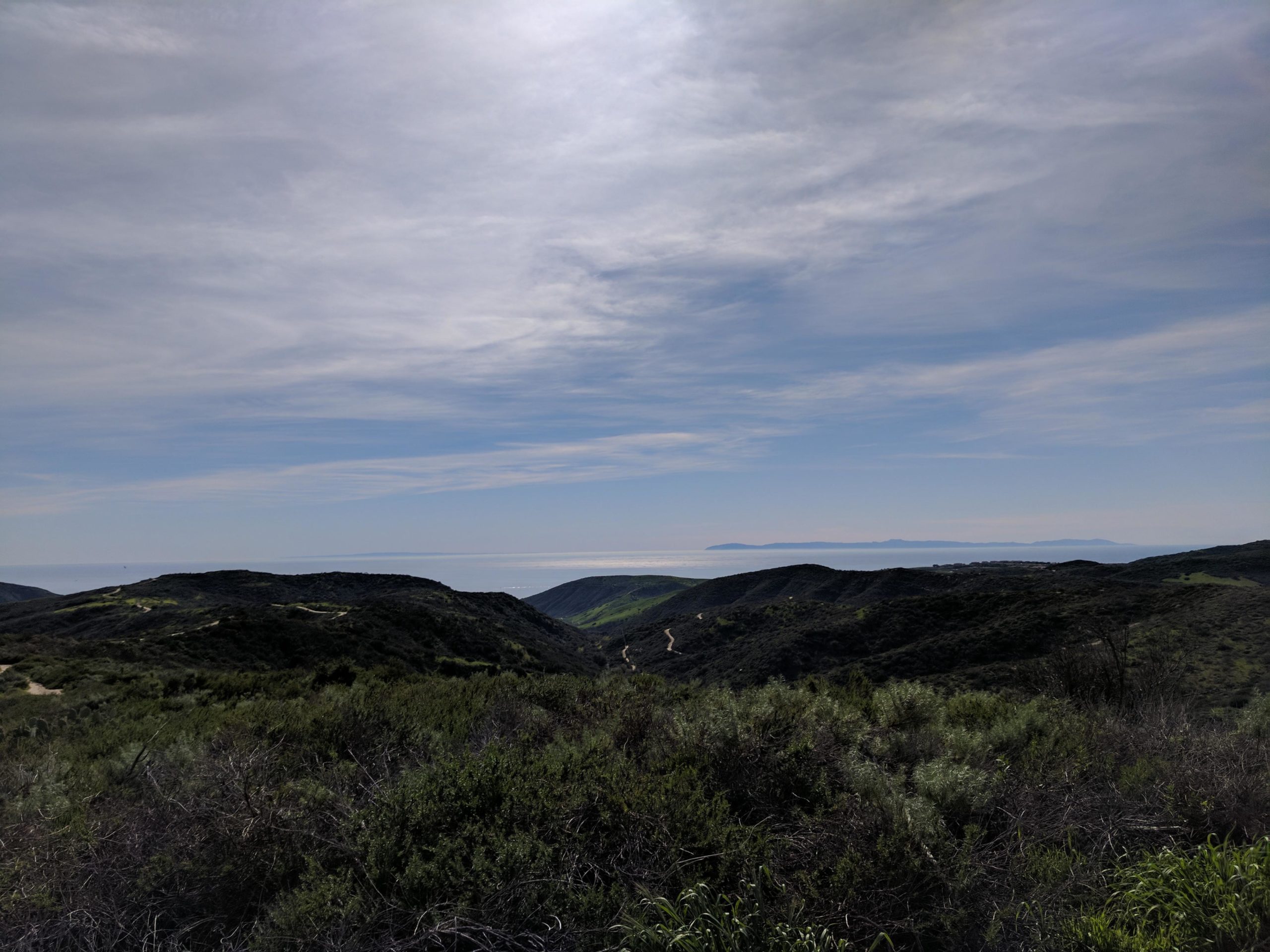 A panoramic view of rolling hills and mountains under a partly cloudy sky, with the ocean visible in the distance. The foreground features lush green vegetation and winding pathways leading through the landscape. El Moro / Erection Loop mountain bike trail.