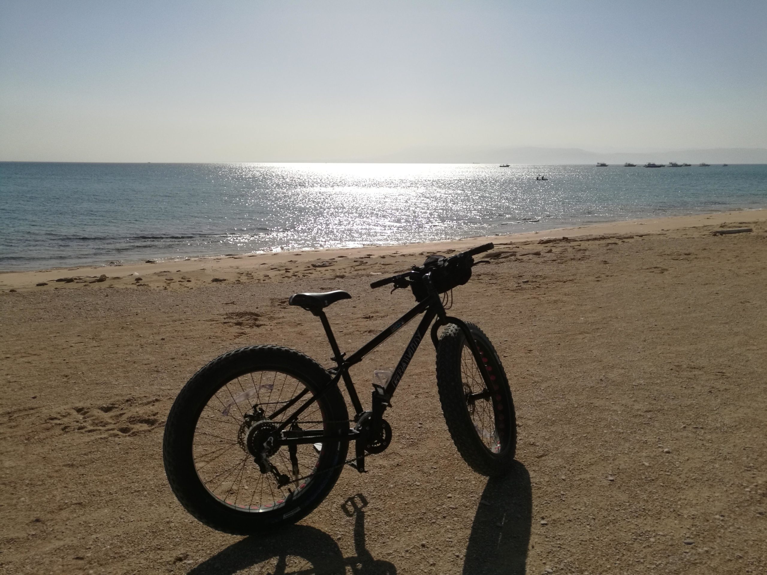 A black fat bike parked on a sandy beach with calm blue water in the background, reflecting sunlight. The scene captures a serene coastal landscape with gentle waves and a few boats in the distance under a clear sky. Matarma Bay Beach mountain bike trail.
