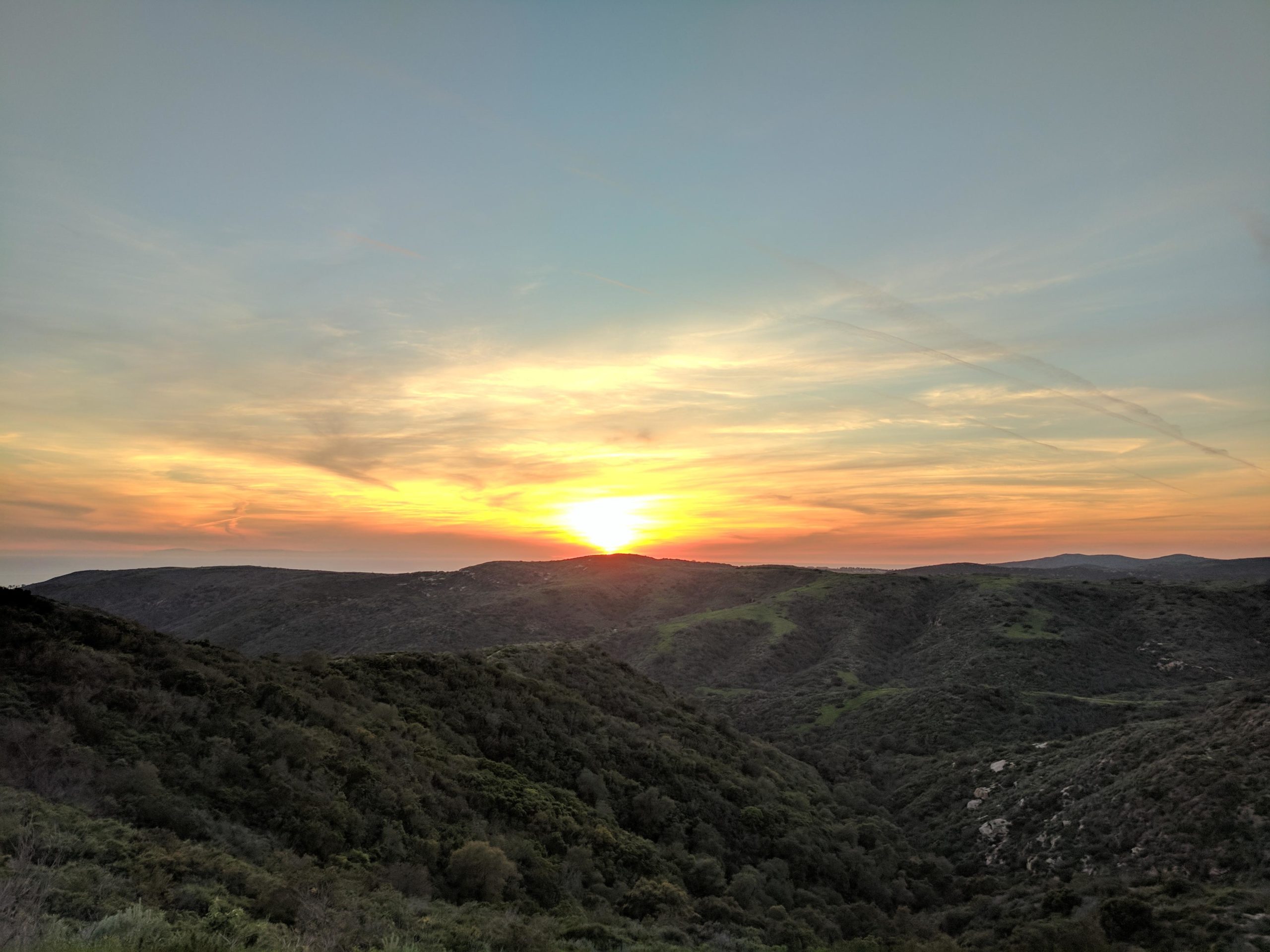 A scenic landscape at sunset, showcasing rolling green hills under a sky painted with warm hues of orange, yellow, and blue. The sun is setting on the horizon, casting a soft glow over the natural surroundings. El Moro / Erection Loop mountain bike trail.
