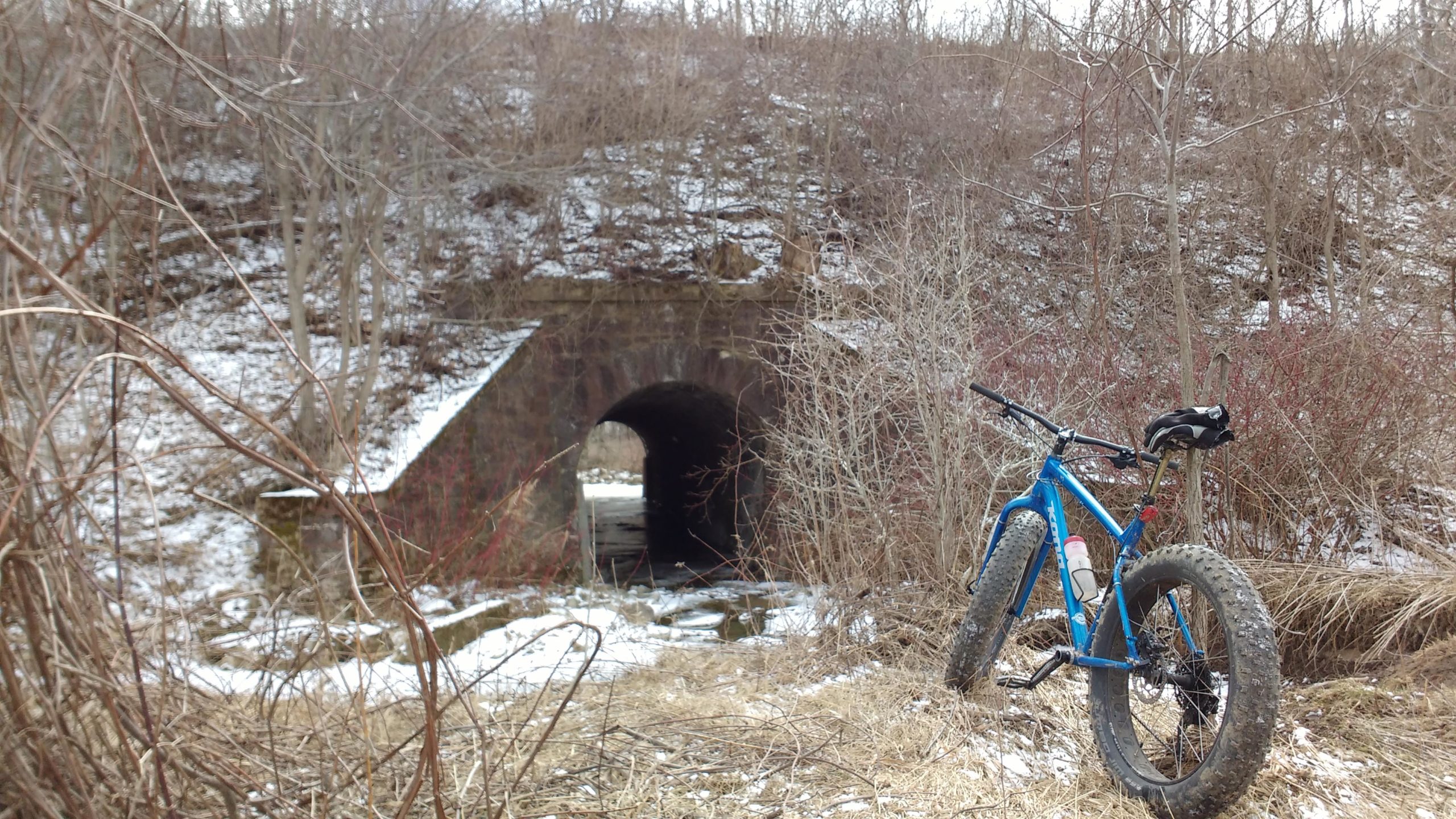 A blue fat tire bike is parked on the grass near a snowy landscape, with a stone tunnel in the background. The area is surrounded by bare trees and shrubs, typical of a winter setting. Lafortune Park mountain bike trail.