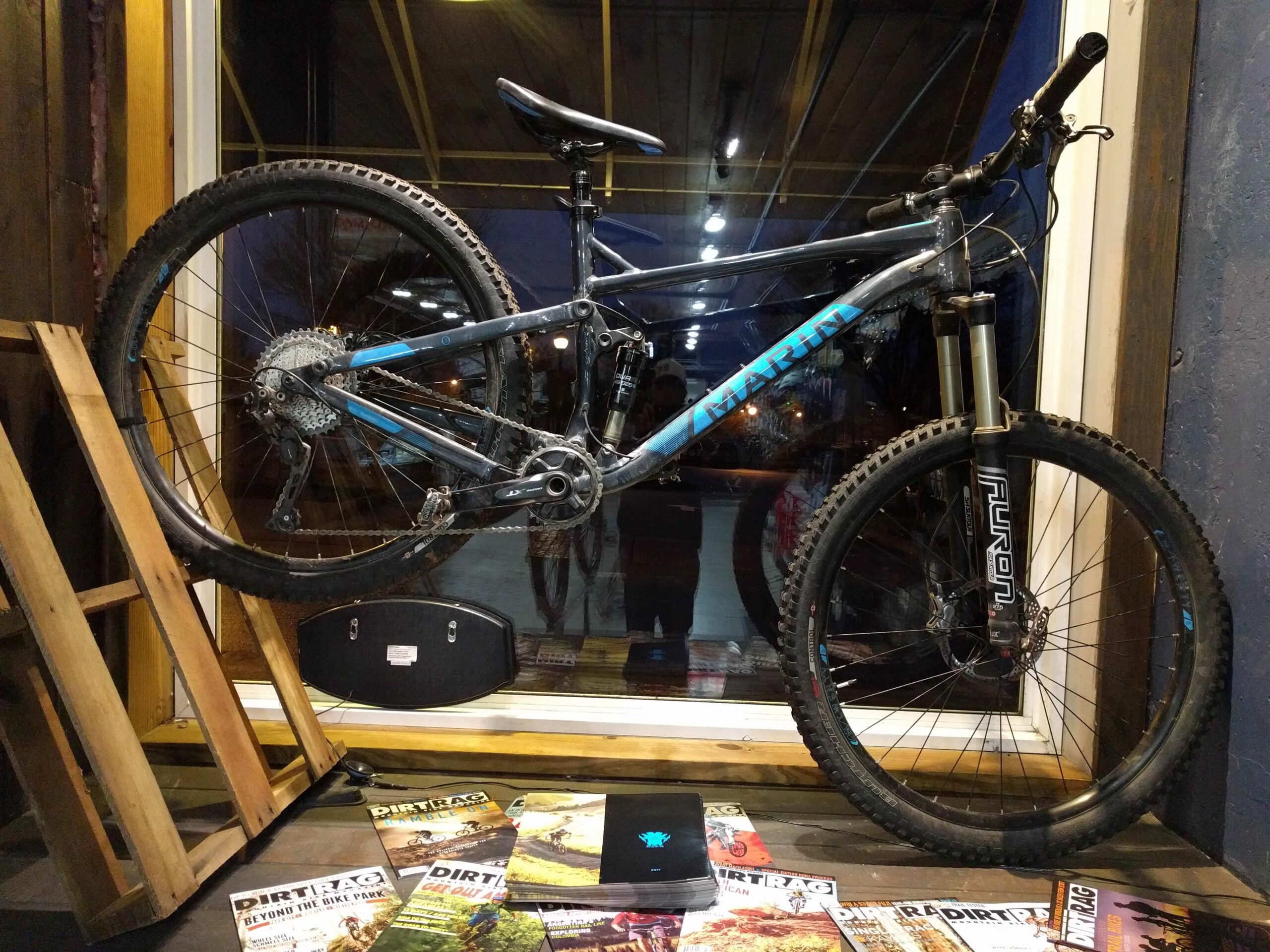 Marin Hawk Hill: A mountain bike displayed against a window, showcasing its frame and components. The bike features black tires with a blue and silver frame, mounted on a wooden display stand. Below the bike, there are several biking magazines spread out on a table. The setting has an inviting atmosphere with soft lighting in the background.