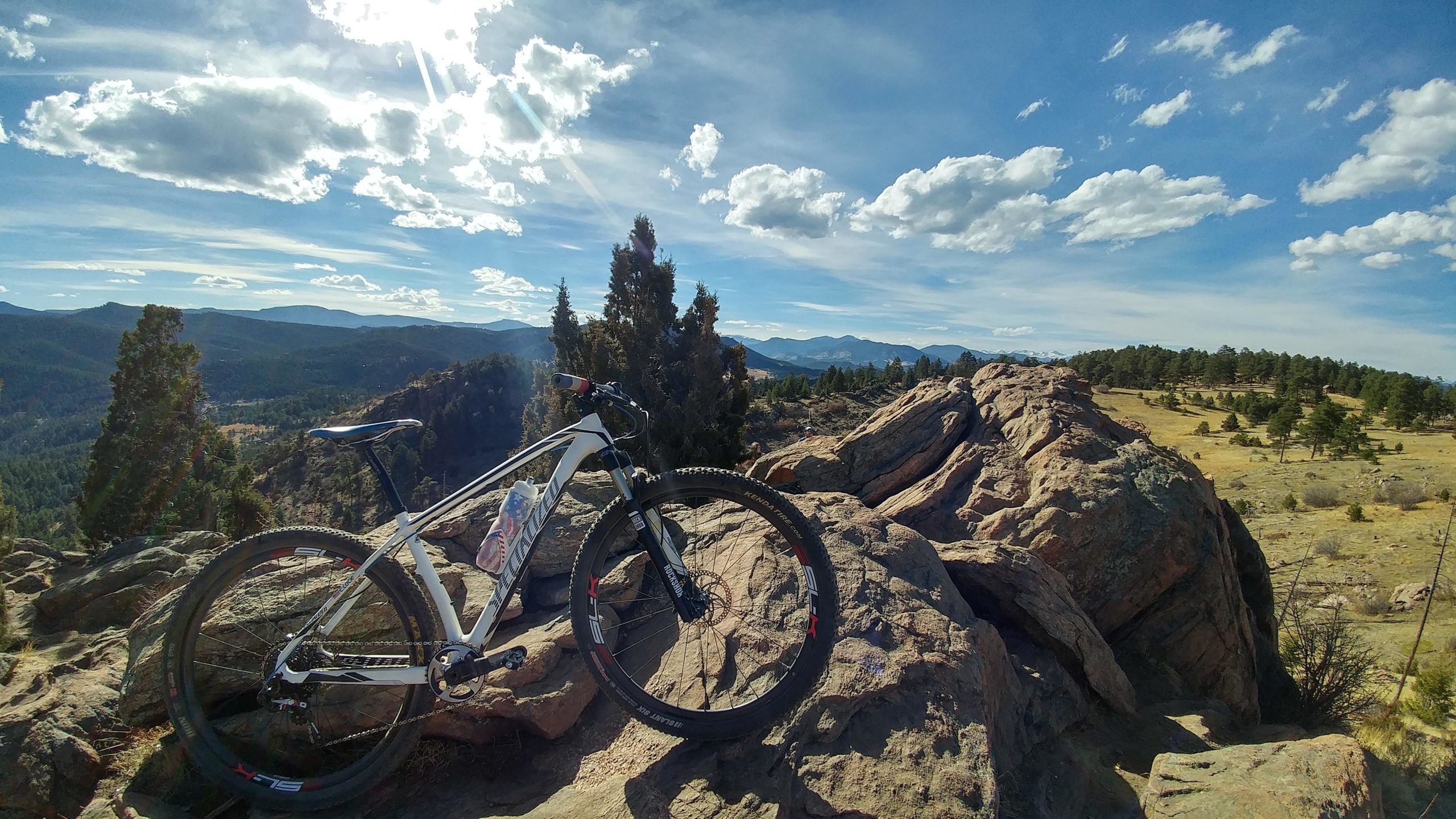 A mountain bike resting on a large rock outcrop, overlooking a vast landscape of rolling hills and trees under a bright blue sky with fluffy clouds. The scene captures a tranquil outdoor setting, ideal for biking and exploring nature. Mount Falcon Park mountain bike trail.