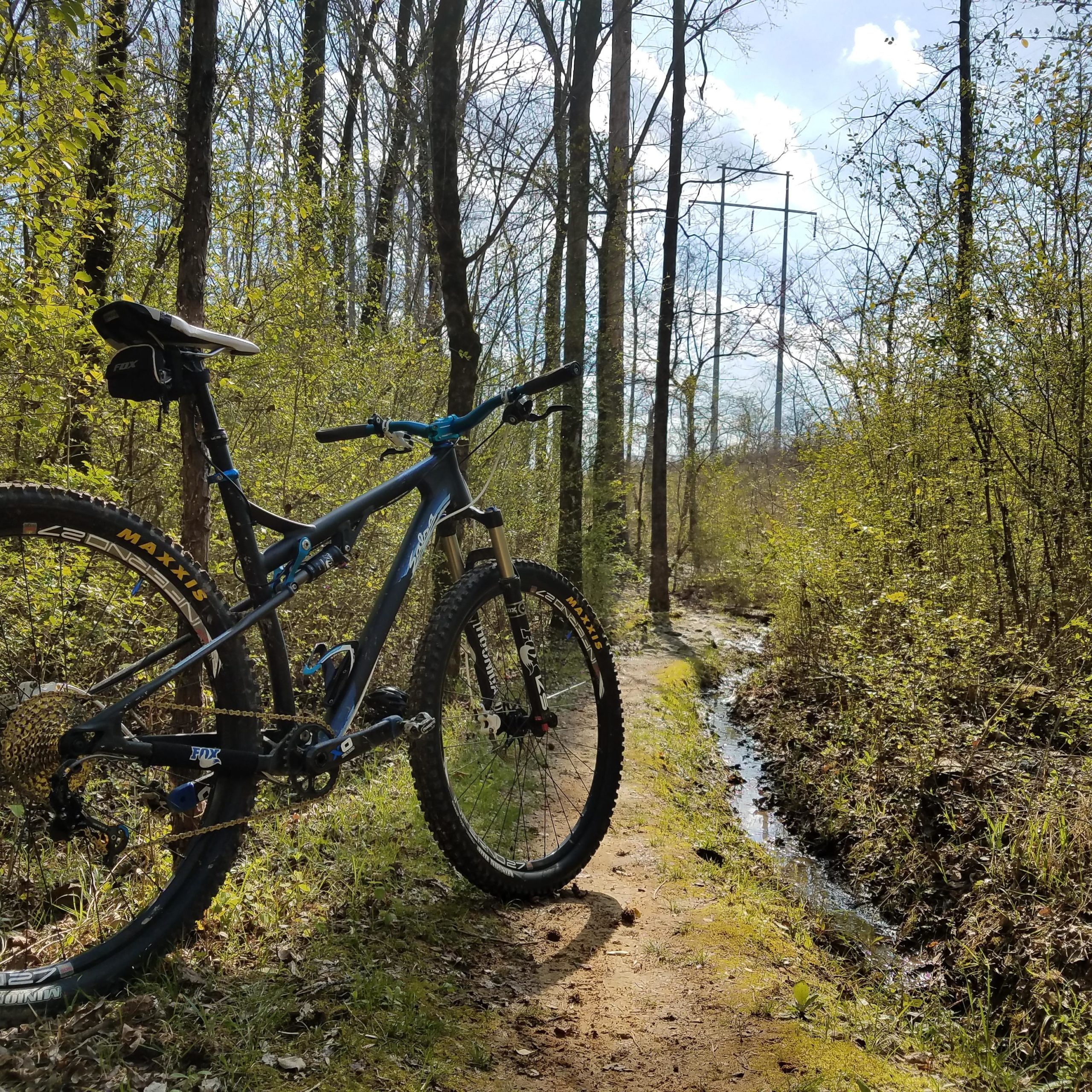 A mountain bike is parked on a dirt trail surrounded by lush greenery and trees, with a small stream running alongside. The sky is partially cloudy, indicating a pleasant day for outdoor activities. Research and Technology Park (ERC) mountain bike trail.