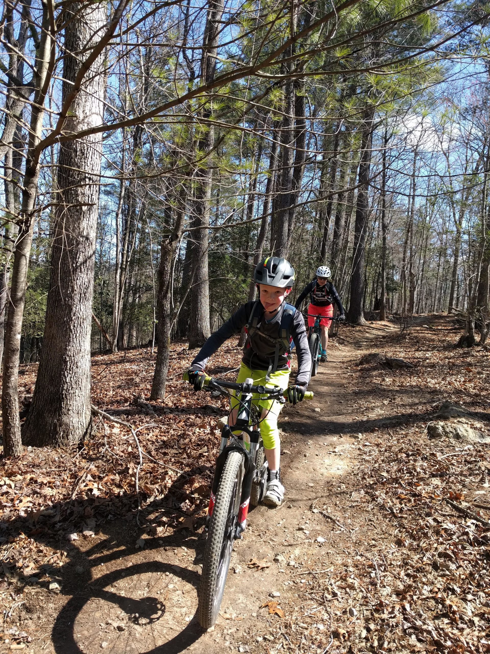 Two children ride mountain bikes along a dirt trail in a forest. The first child is in bright yellow biking gear and a helmet, while the second is further back and wearing red shorts. Surrounding them are tall trees and a clear blue sky. Fallen leaves cover the ground, indicating early spring or late fall. Warrior Creek mountain bike trail.