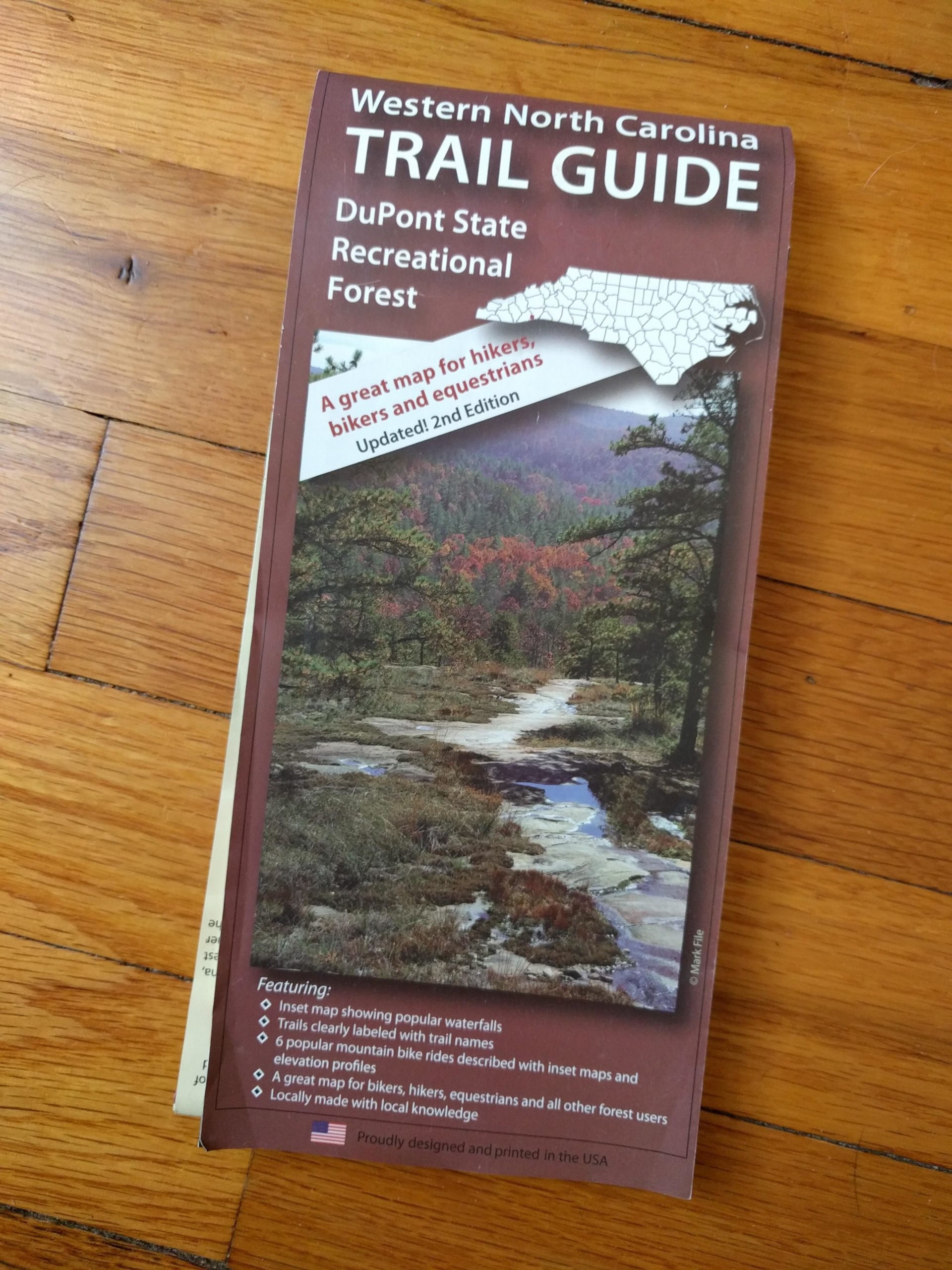 Alt text: "Cover of the 'Western North Carolina Trail Guide' for DuPont State Recreational Forest, featuring a scenic image of a waterway and forest. The guide highlights trails for hikers, bikers, and equestrians, with notes on maps, elevation profiles, and local knowledge." DuPont State Forest mountain bike trail.
